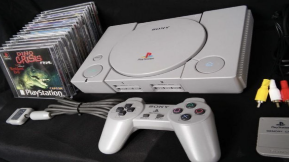 A Sony PlayStation console with a controller, memory card, AV cables, and a stack of game cases&mdash; including &ldquo;Dino Crisis&rdquo;&mdash; arranged on a black surface, showcases one of the most iconic consoles and its essential accessories.