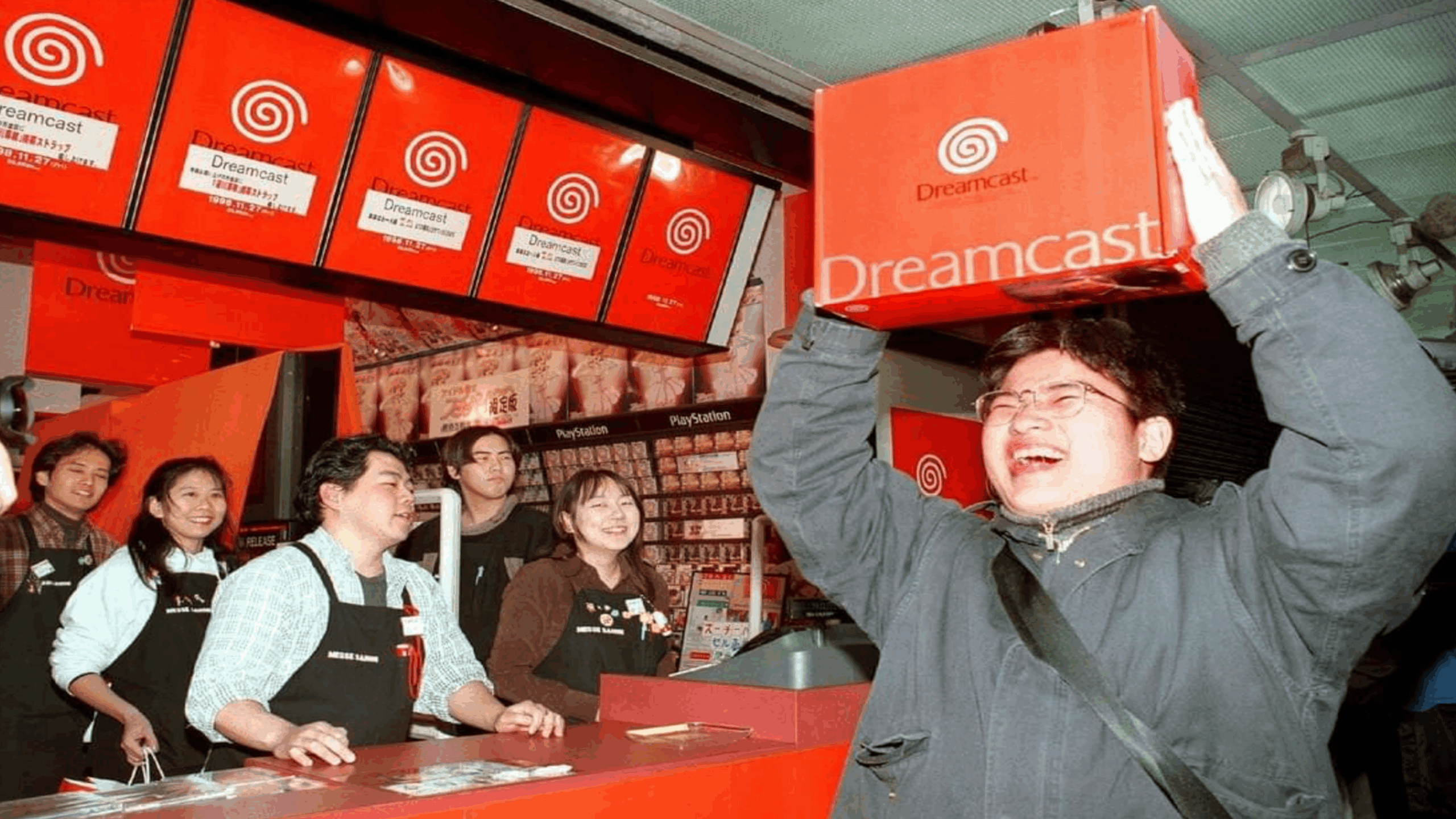 A man excitedly holds a boxed Sega Dreamcast console above his head in a store, while employees and other customers smile and watch him.