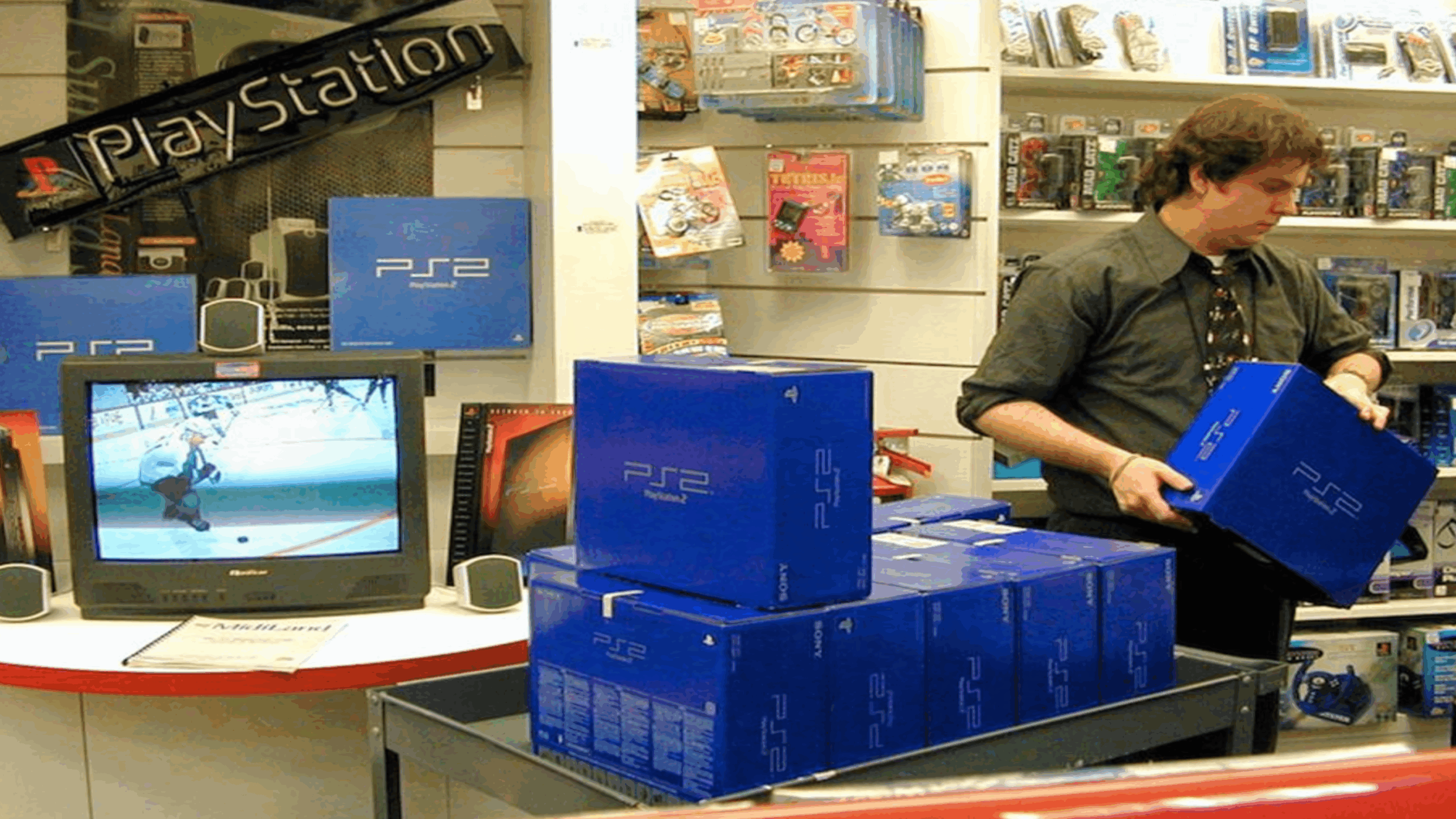 A store employee stacks blue PlayStation 2 consoles near a TV displaying a hockey game, with PlayStation accessories on shelves in the background.