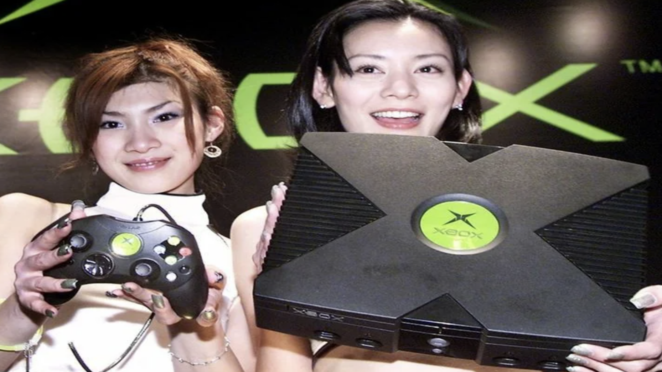 Two women smiling at a gaming event, one holding an original Xbox console and the other holding an Xbox controller, surrounded by a vibrant display of classic consoles, with a large Xbox logo in the background.