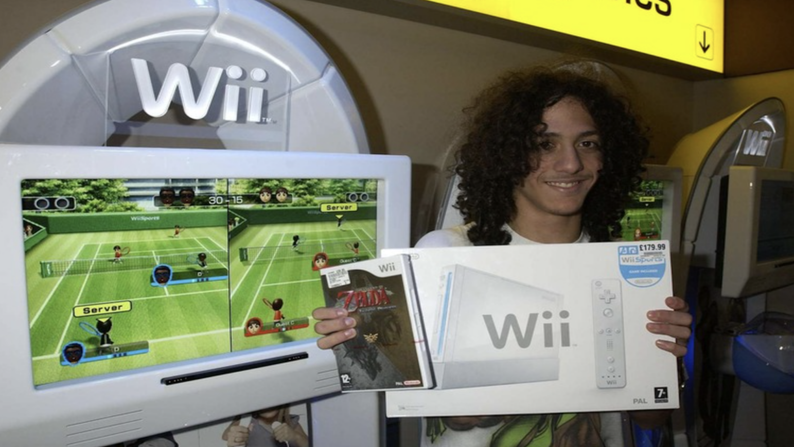 A person with curly hair smiles while holding a Nintendo Wii console box and a Wii game case in front of a display playing Wii Sports tennis, showcasing their excitement for classic gaming consoles.