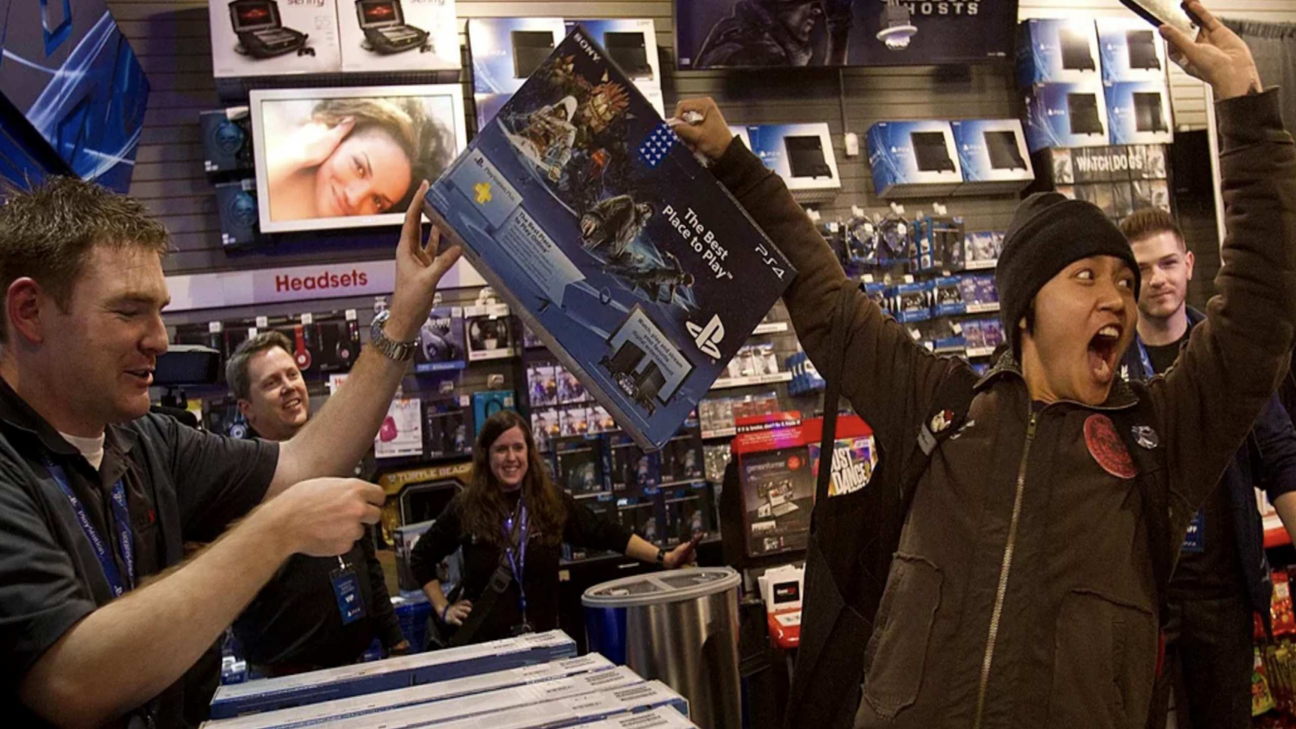 A joyful person holds up a PlayStation 3 console box in a store as employees and other customers cheer, surrounded by video game consoles, merchandise, and electronics.