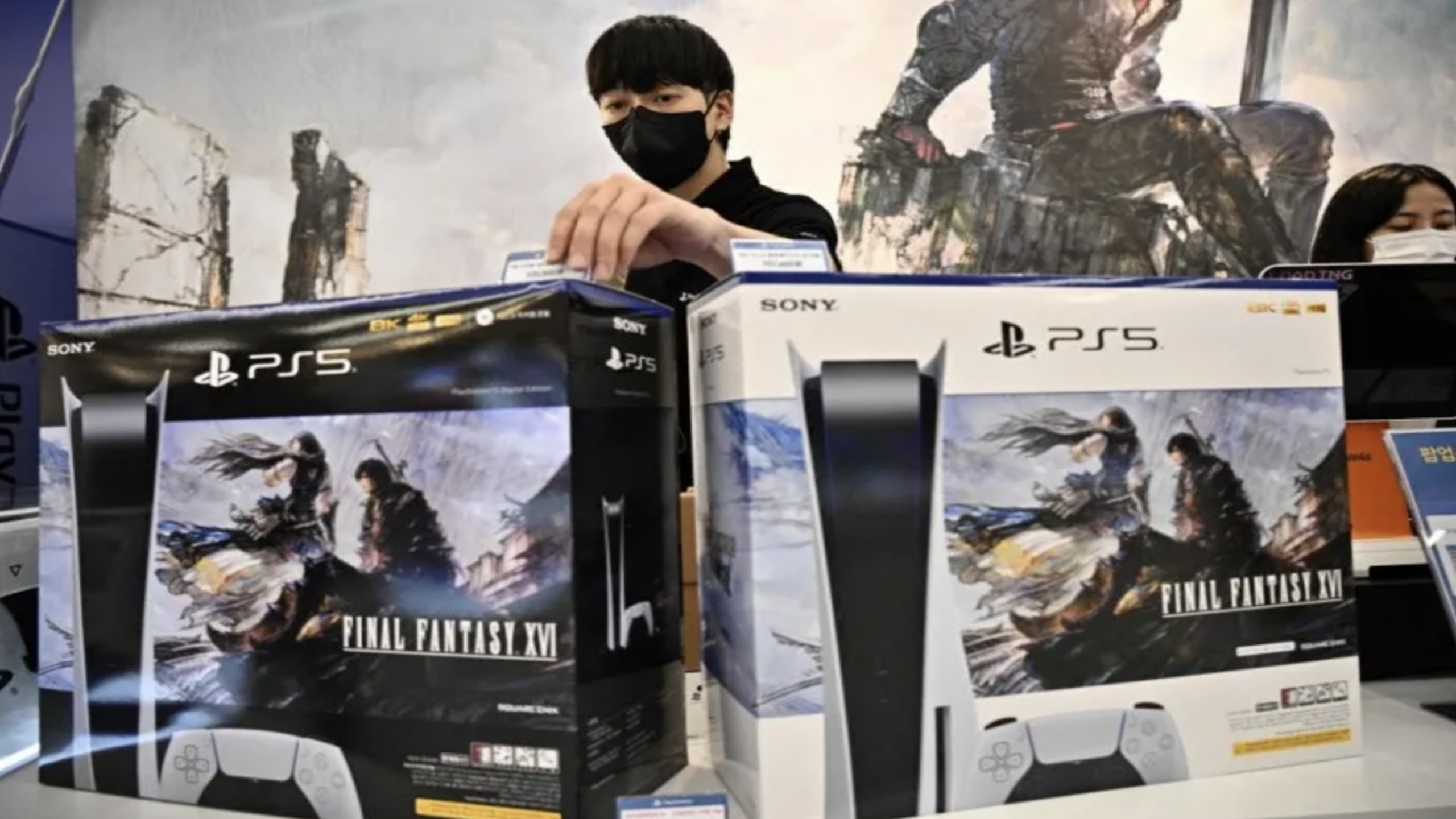A person wearing a mask arranges PlayStation 5 consoles with Final Fantasy XVI artwork on the boxes at a store counter. Another masked person is visible in the background, surrounded by stacks of coveted gaming consoles.