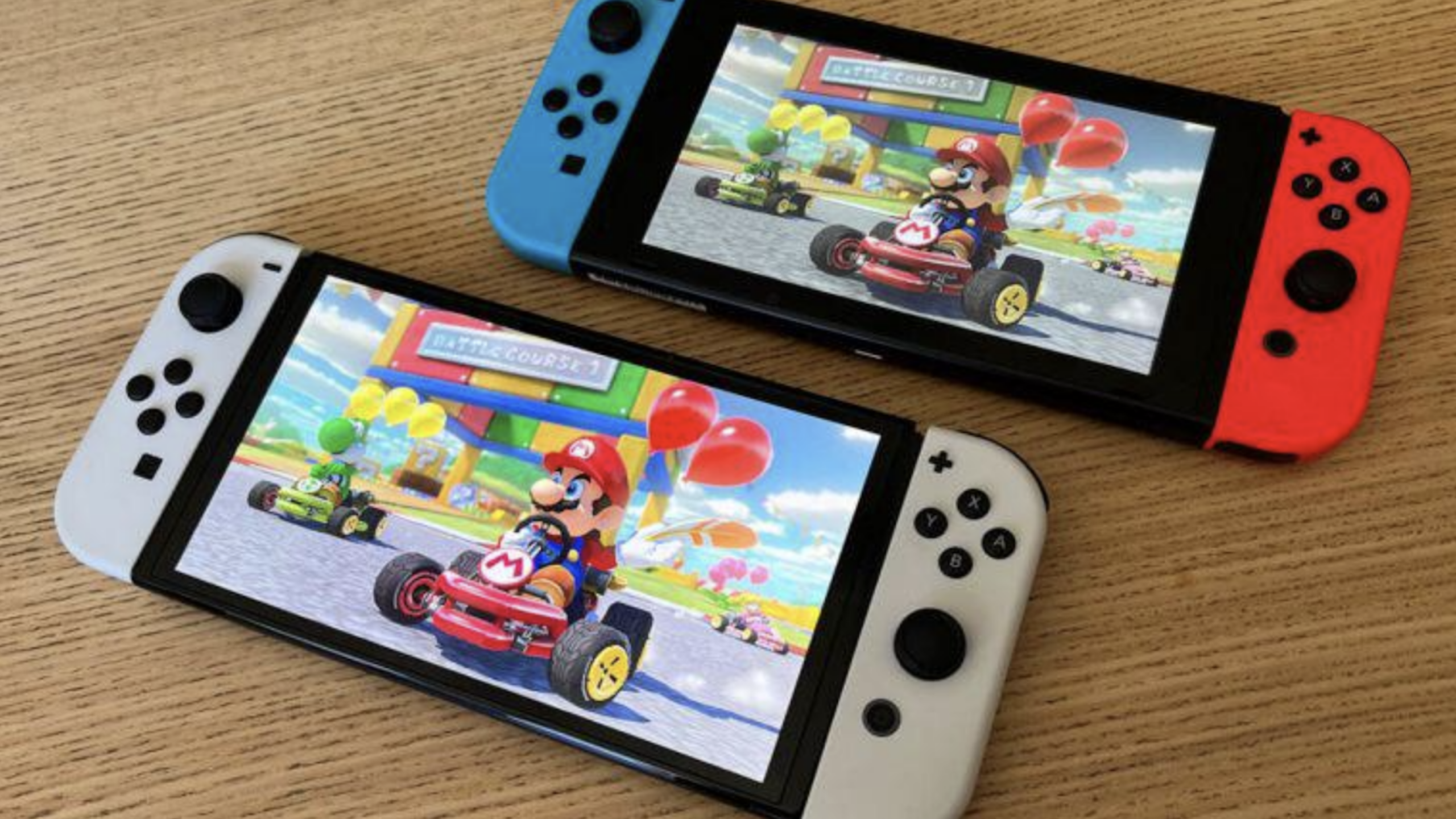 Two Nintendo Switch consoles rest on a wooden surface, each displaying Mario Kart gameplay featuring Mario in a go-kart with balloons attached, racing near a colorful building. The vibrant screens highlight the fun and dynamic experience these consoles deliver.
