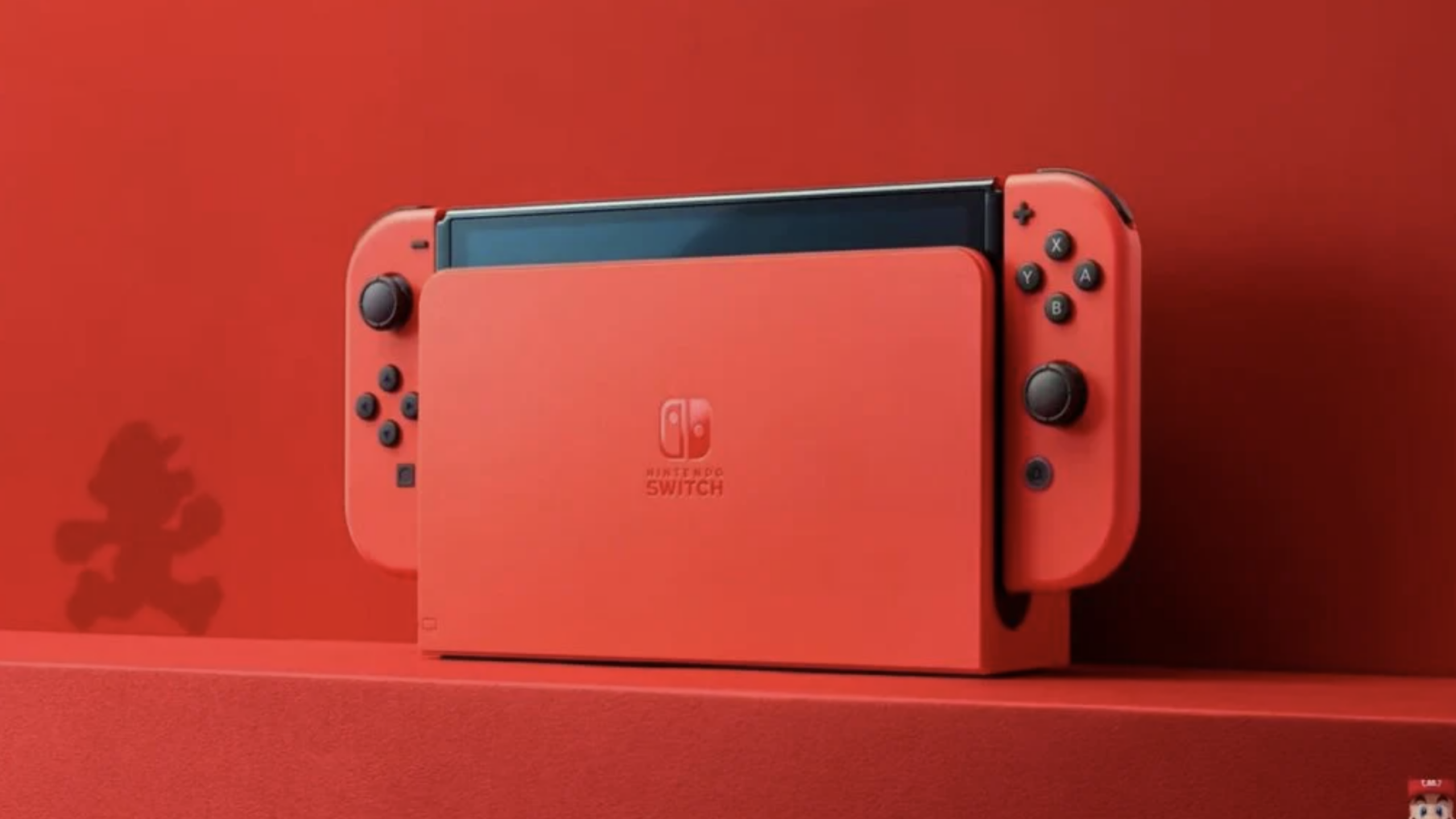 A red Nintendo Switch console with Joy-Con controllers is docked in a matching red dock, showcasing the elegance of Nintendo consoles. The background and surface are also red, with a subtle silhouette of Mario visible on the left.