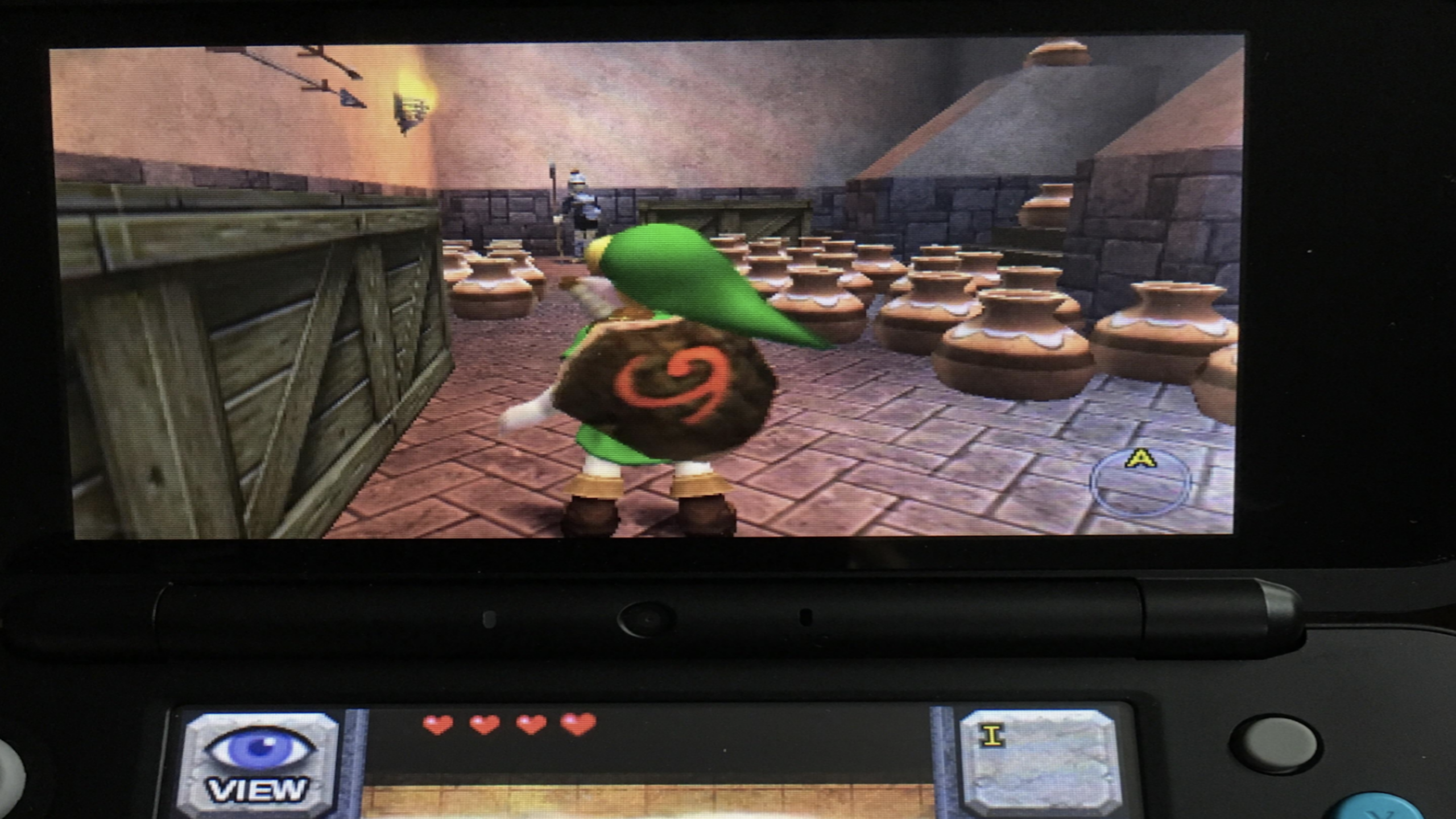 A video game scene on a handheld console shows a character in green, Link, standing in a room filled with pots and crates in "The Legend of Zelda: Ocarina of Time 3D." This classic video game moment captures the spirit of exploration and adventure fans love.