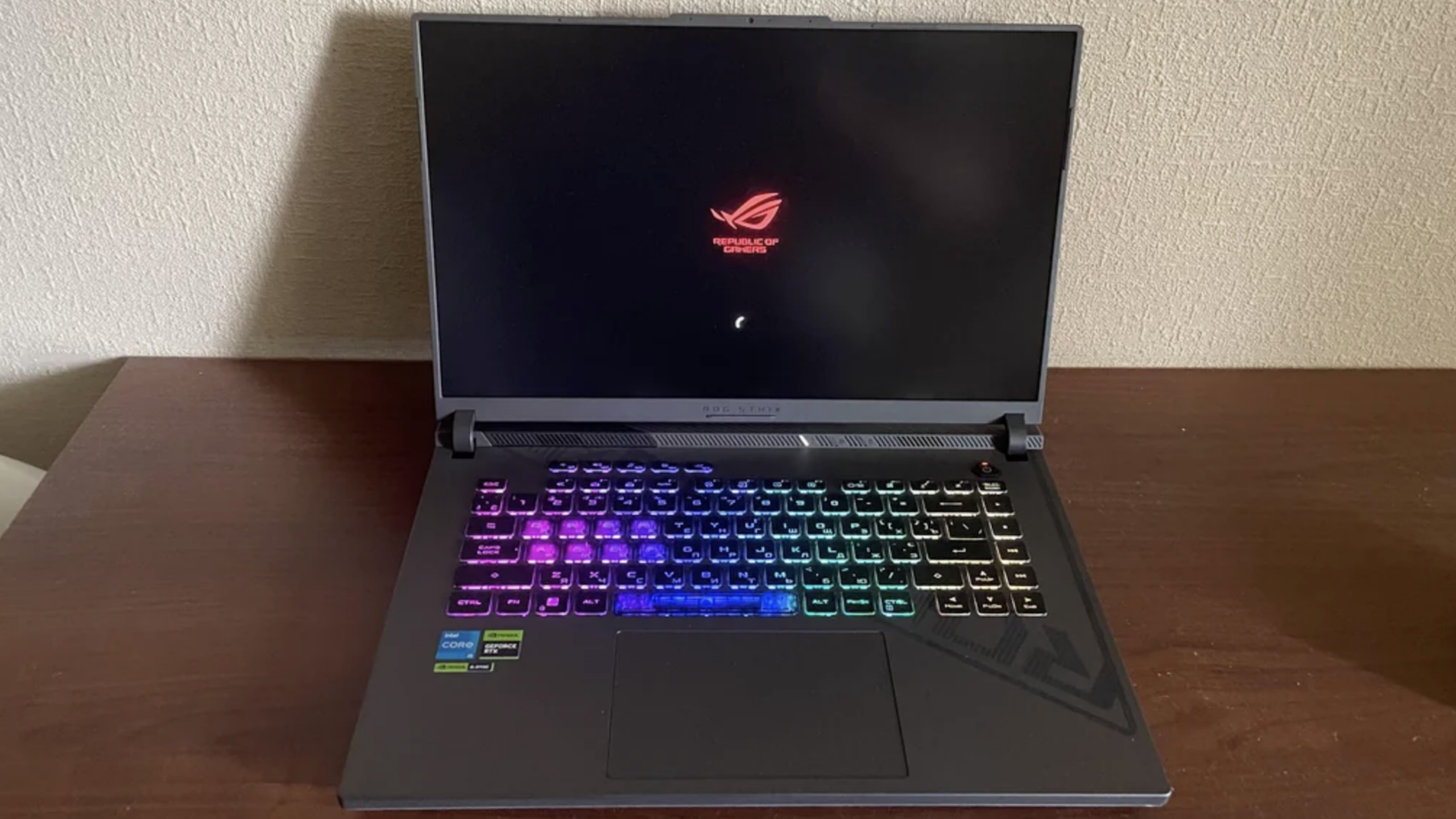 A black ASUS ROG laptop sits on a wooden surface, displaying the startup screen. Its keyboard is illuminated with rainbow RGB lighting, evoking the vibrant aesthetic often seen in gaming consoles.