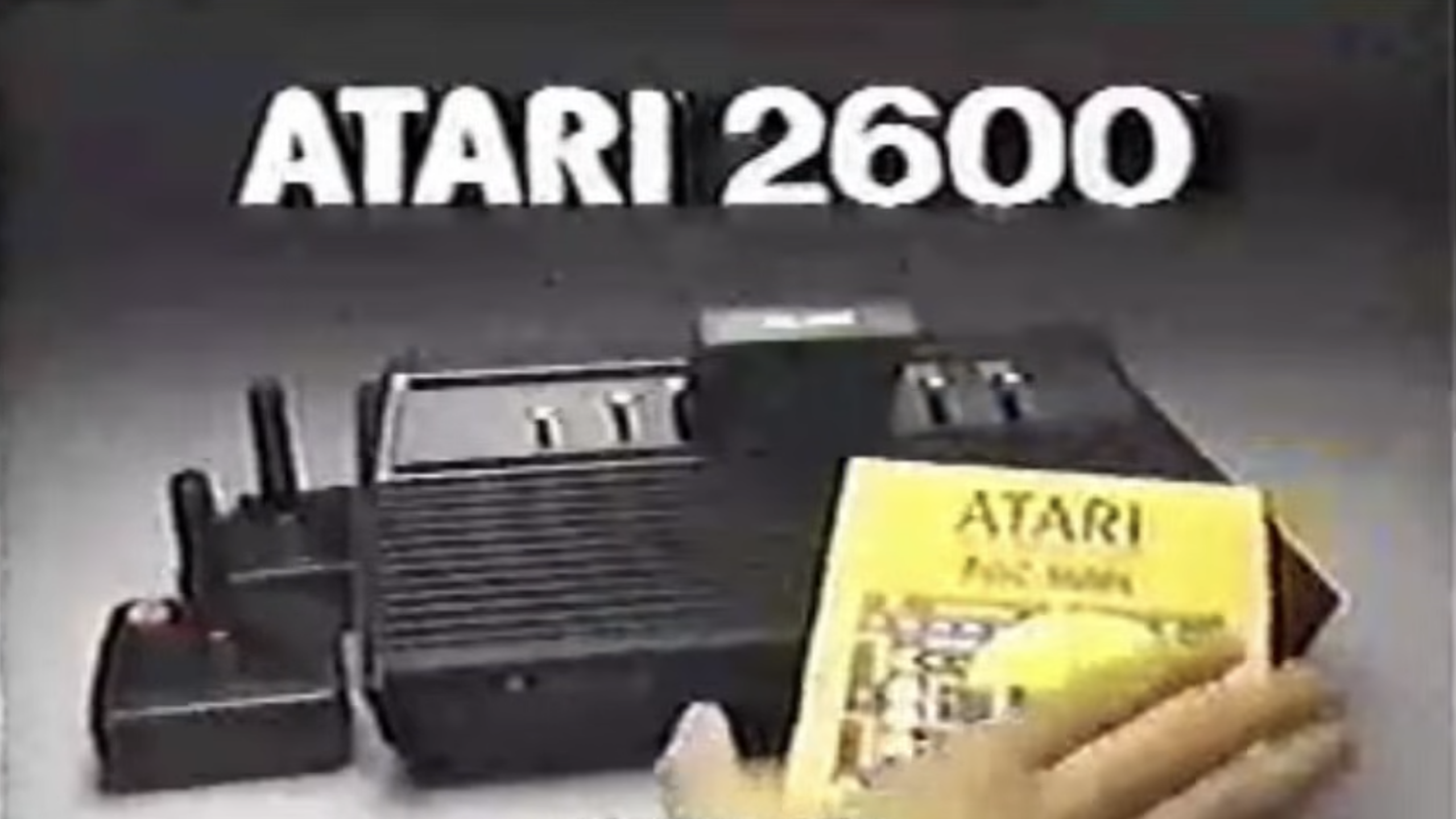 A close-up of an Atari 2600 console with two joysticks and a person holding an Atari instruction manual. Large text above reads “ATARI 2600,” showcasing one of the most iconic consoles in gaming history.