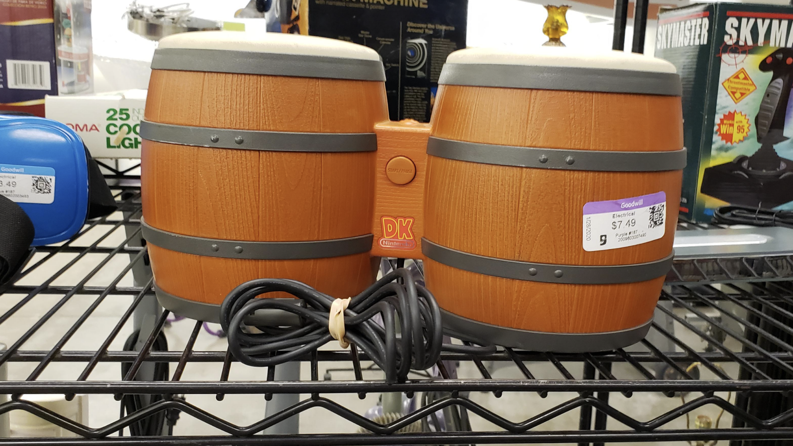 A pair of Donkey Konga bongo controllers with a cord, displayed on a metal shelf in a store alongside other gaming accessories, with price stickers attached.
