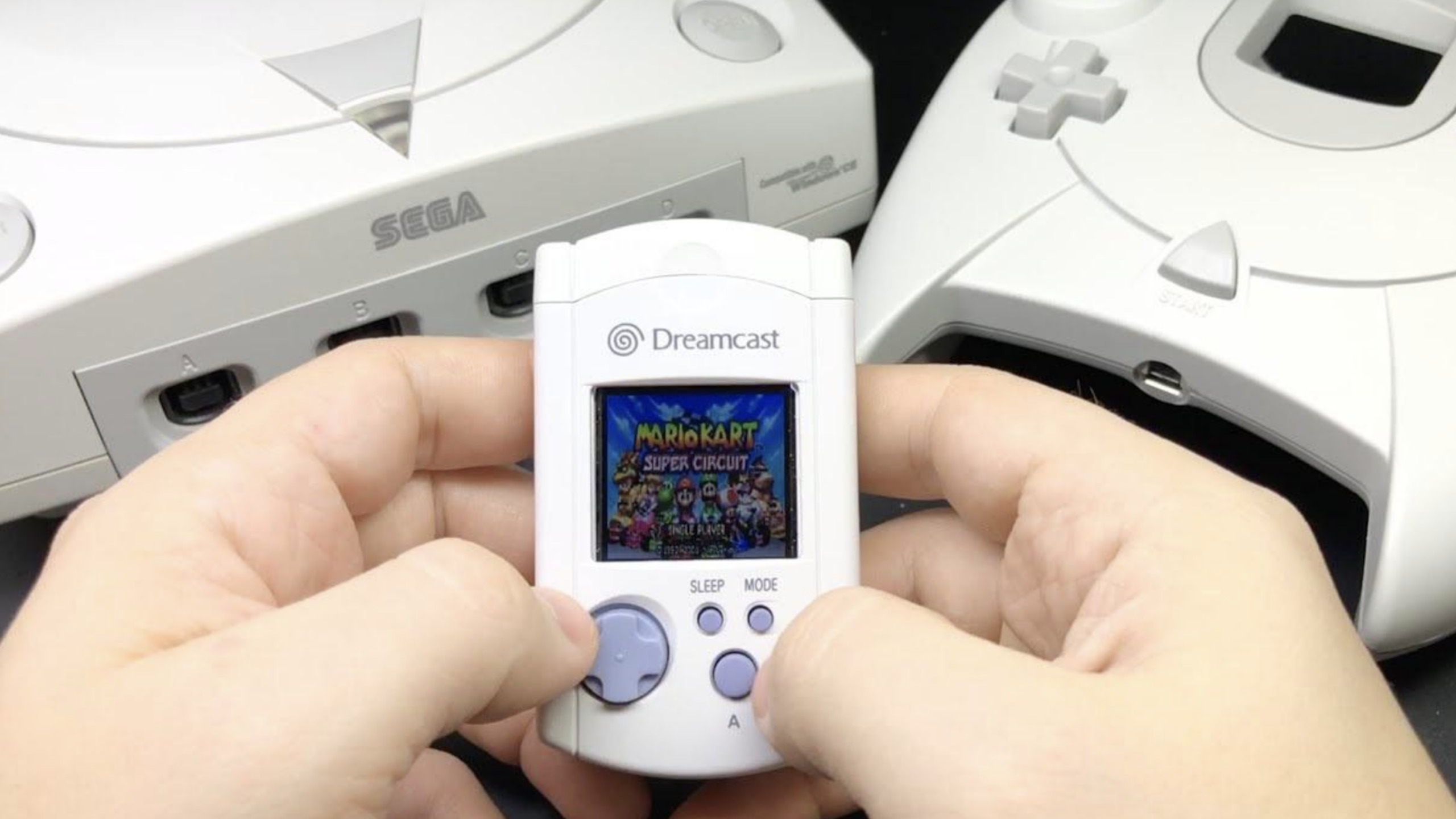 Two hands hold a white Sega Dreamcast VMU, a classic piece among gaming accessories, displaying "Mario Kart Super Circuit" on its screen, with Sega Dreamcast consoles visible in the background.
