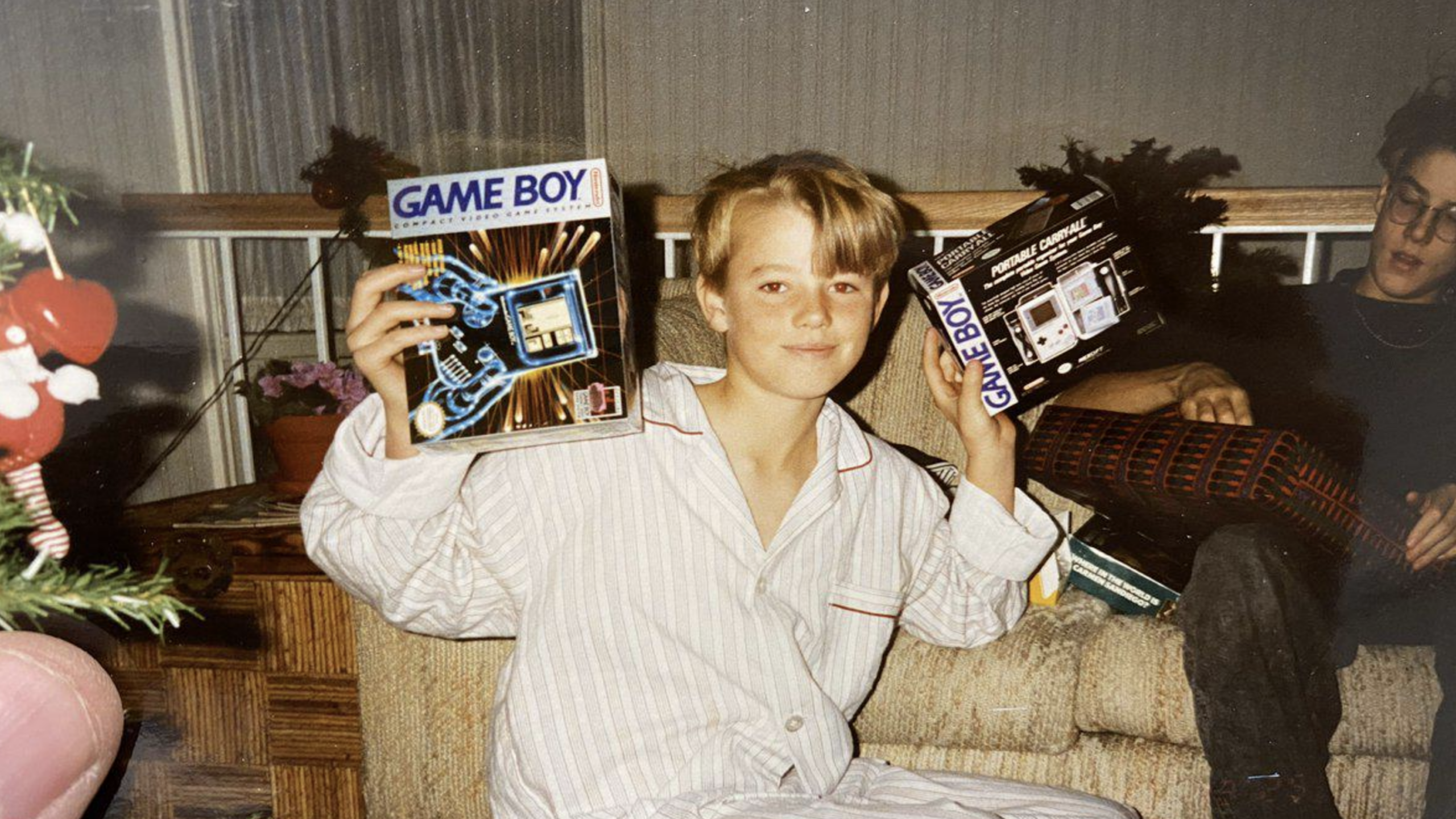 A smiling child in pajamas sits on a couch, holding up a boxed Game Boy and its packaging—one of the iconic Nintendo Consoles—possibly after receiving it as a gift. Another person is seated beside them. Holiday decorations are visible in the corner.