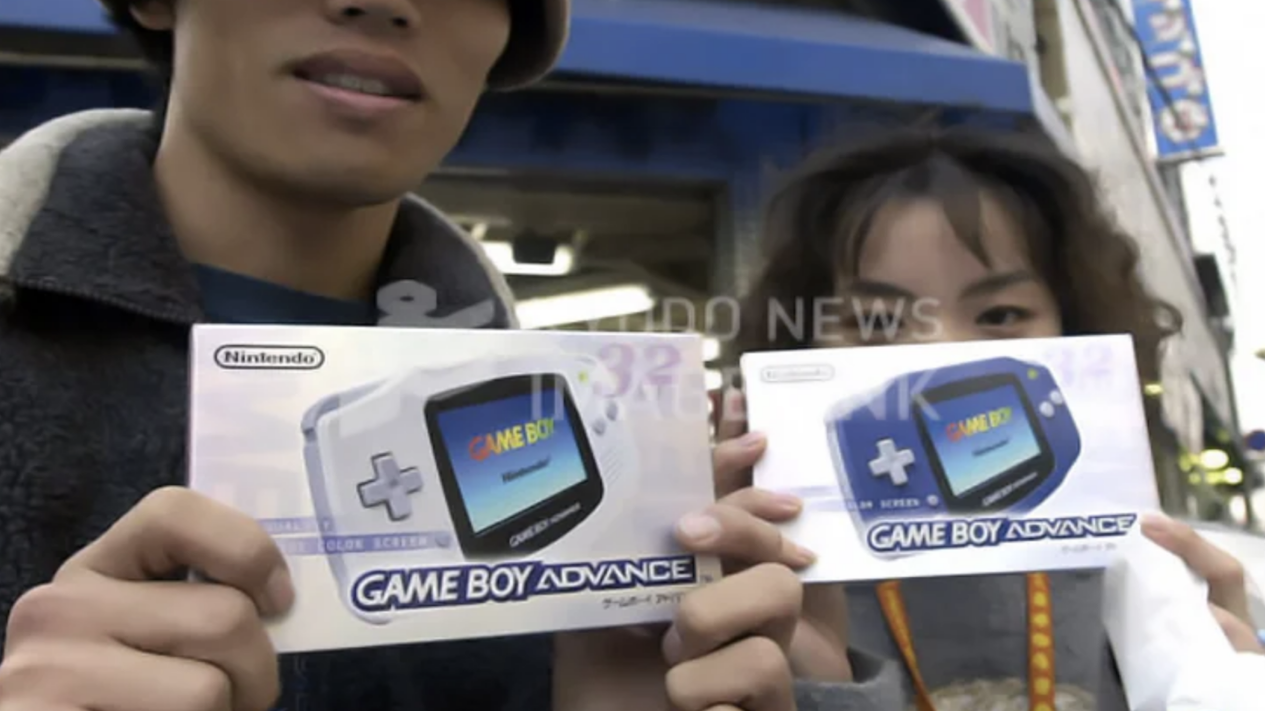 Two people standing outside hold up boxes of Nintendo consoles, specifically Nintendo Game Boy Advance consoles, partially covering their faces. A blue building and blurred signs are visible in the background.
