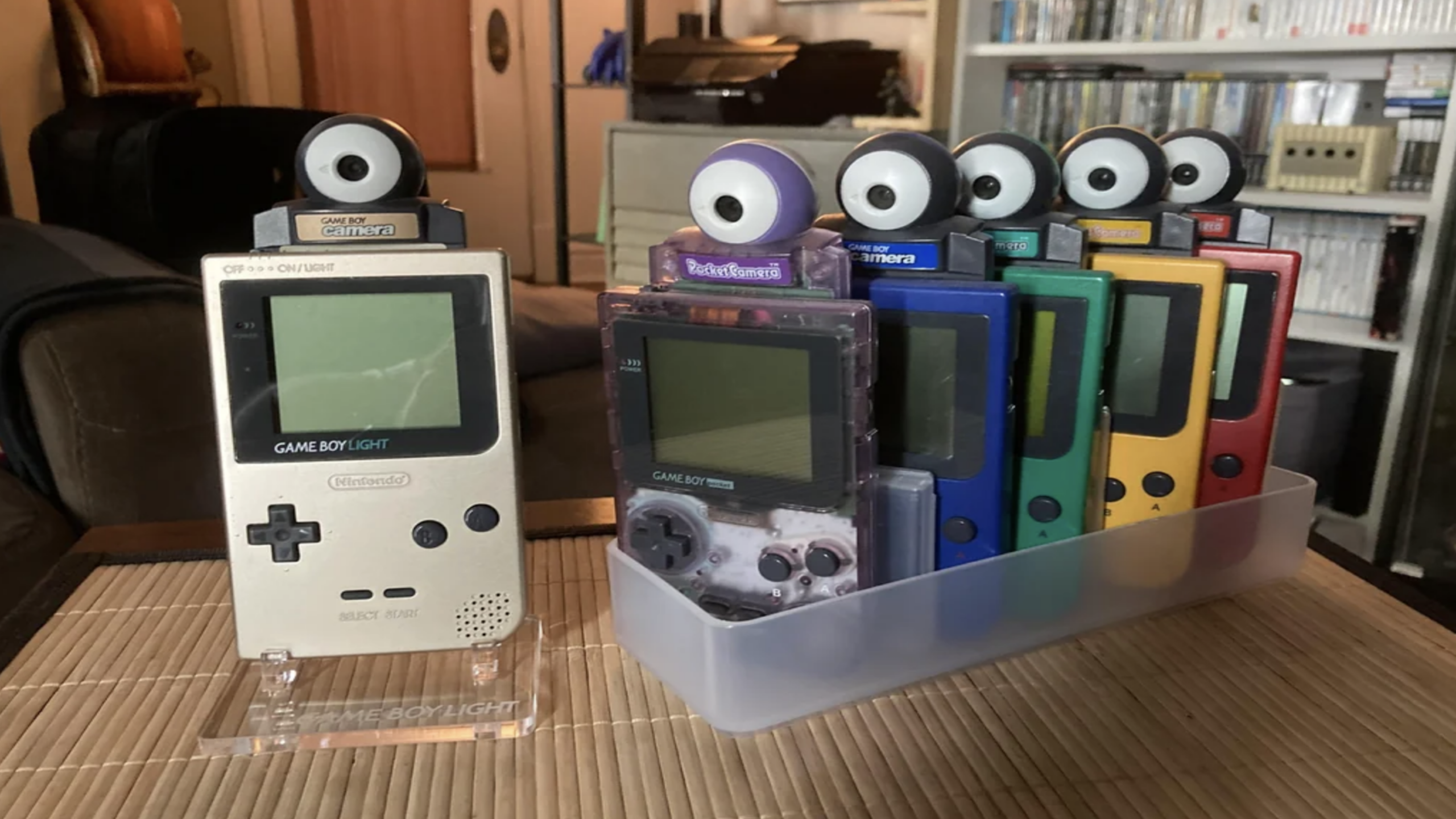 Six Game Boy handheld consoles, each paired with a Game Boy Camera and other classic gaming accessories, are displayed in a row on a table. The consoles come in various colors, including white, purple, green, blue, yellow, and red.