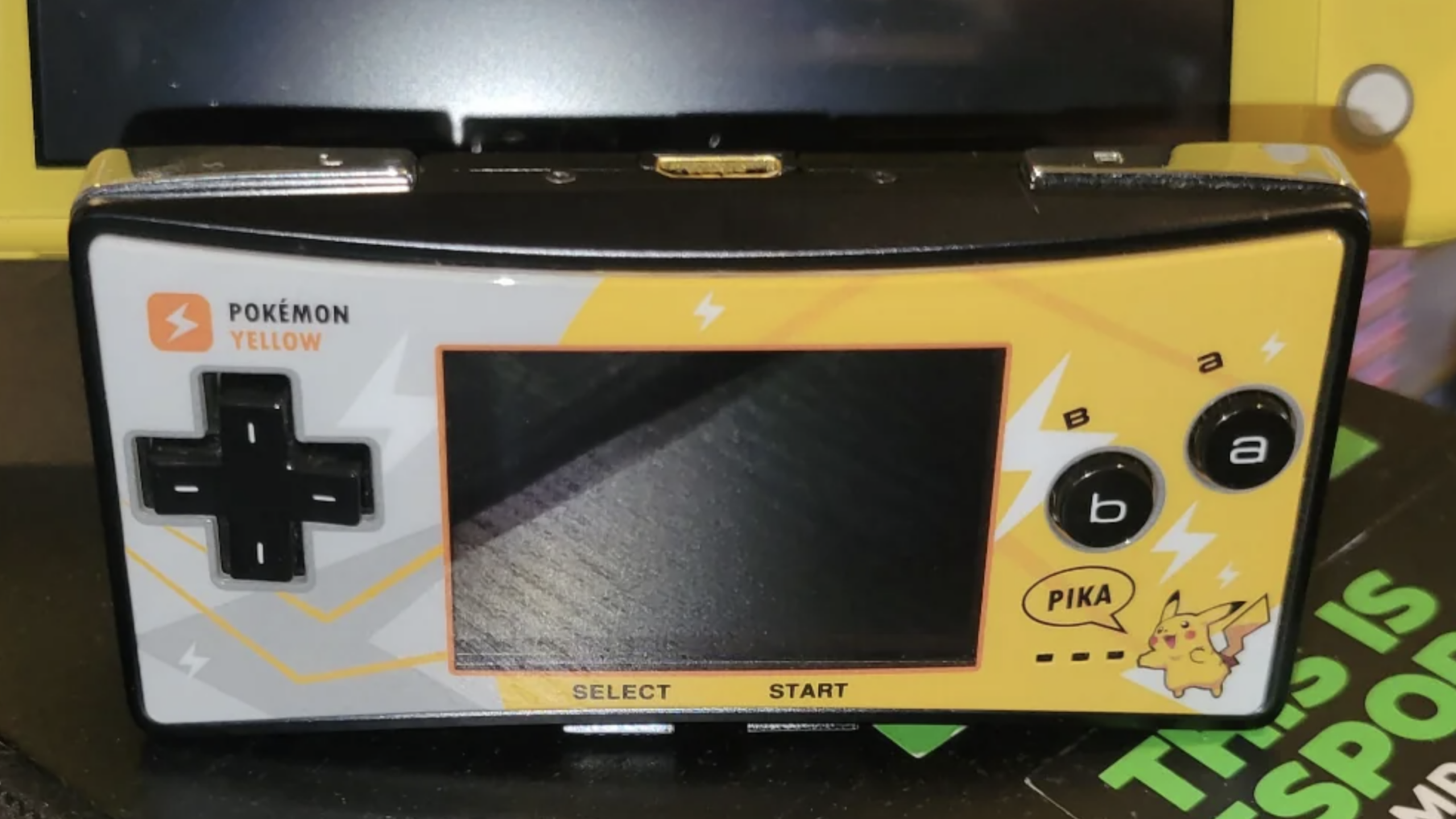 A Game Boy Micro, one of the iconic Nintendo Consoles, with a Pokémon Yellow Pikachu faceplate, showing the screen, control pad, and buttons, resting on a black and green surface.