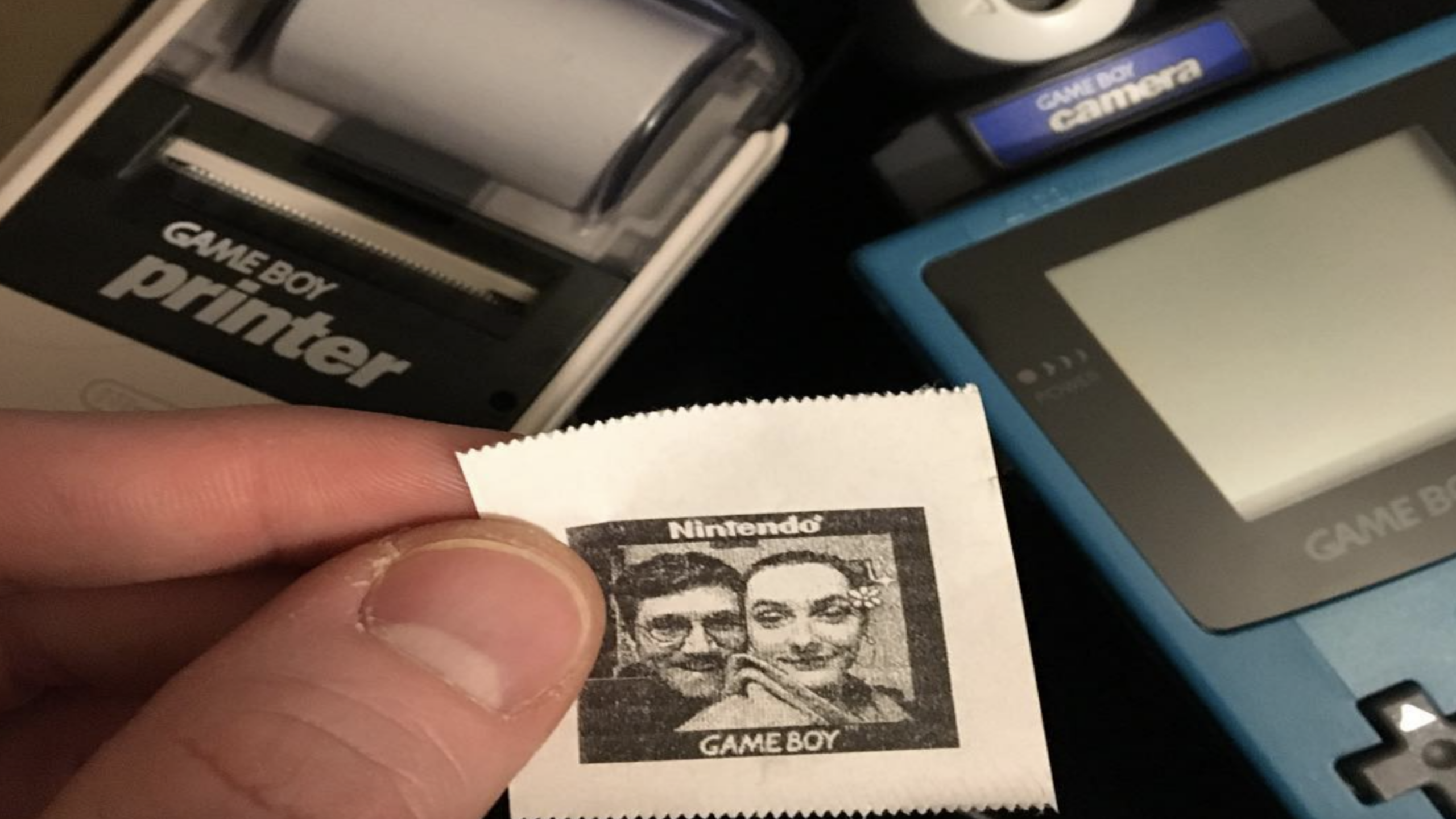A hand holds a small Game Boy Printer photo of two people, with classic gaming accessories like a Game Boy Printer, Game Boy Camera, and a blue Game Boy in the background.