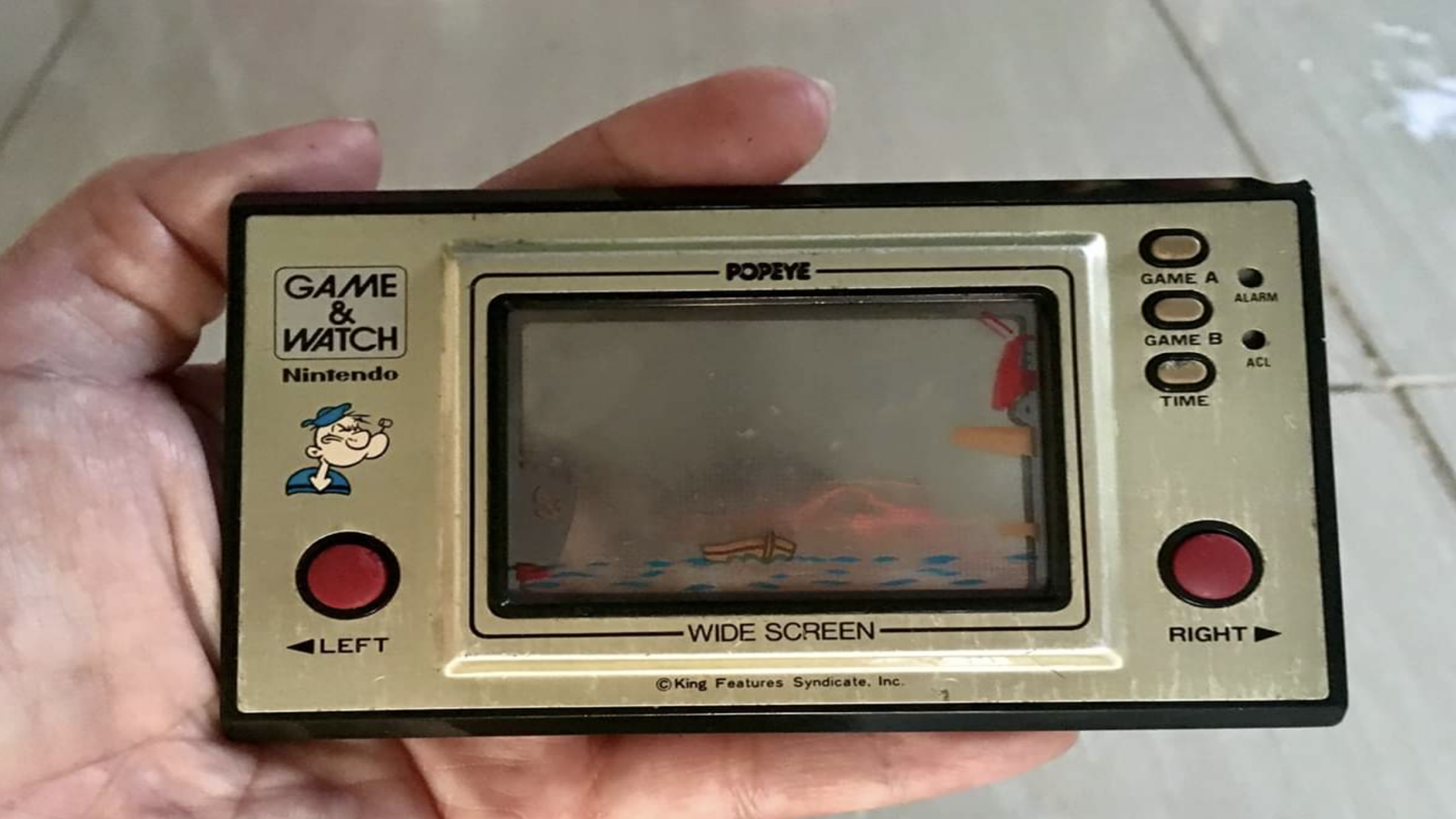 A hand holds a vintage Nintendo Game & Watch "Popeye" handheld device, a nostalgic gem among Nintendo Consoles, featuring two red buttons and a faded screen.