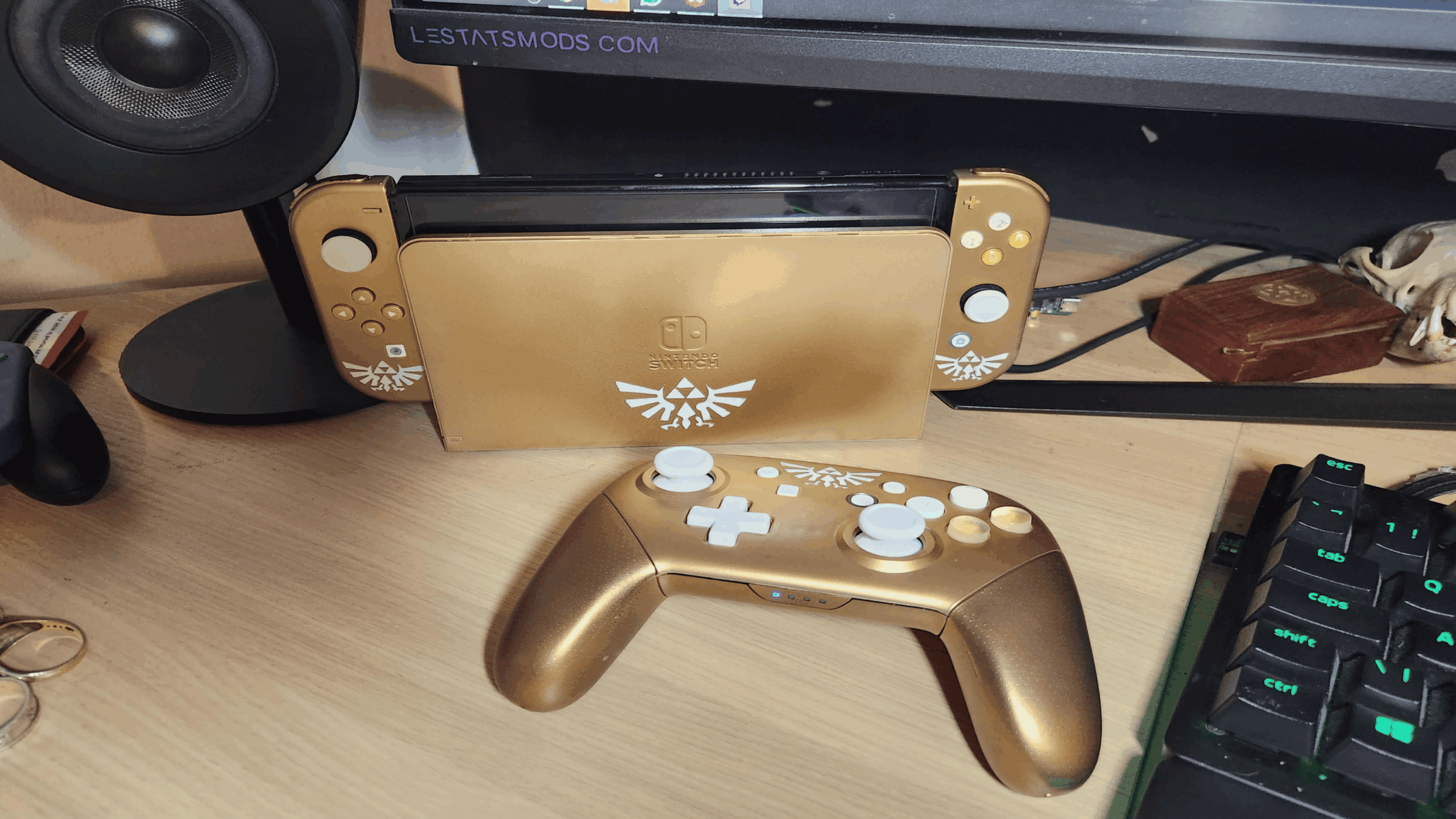 A customized gold Nintendo Switch, one of the most striking gaming consoles, with white buttons and a Legend of Zelda logo sits on a desk. A matching gold and white controller is in front, near a keyboard and computer monitor.