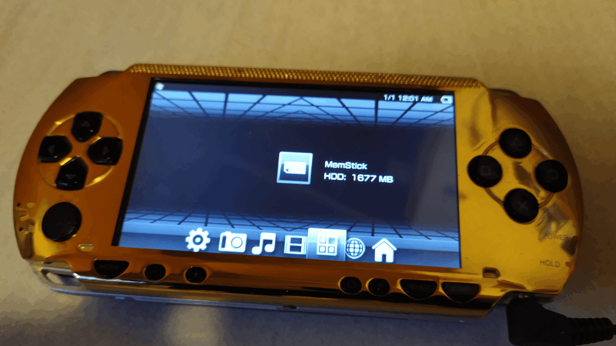 A gold PlayStation Portable (PSP) handheld console, one of the classic gaming consoles, displays its main menu screen with "MemStick" showing 1677 MB available. The background features a blue grid pattern.