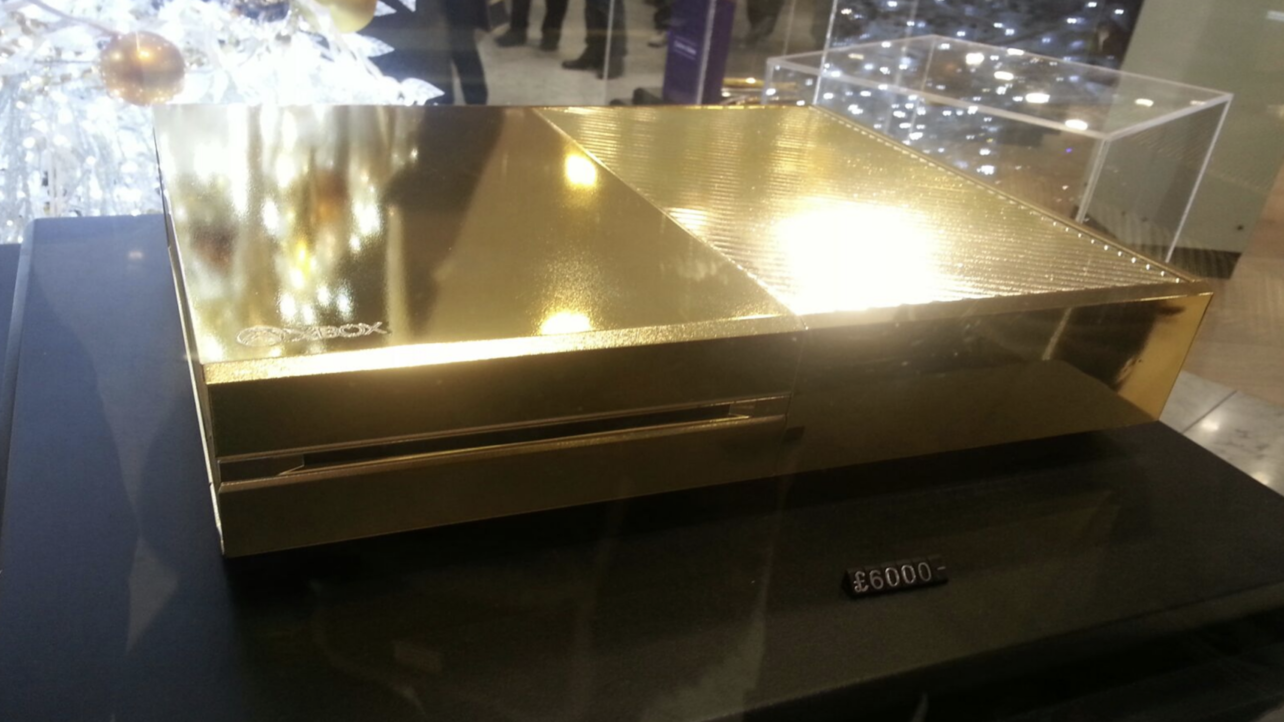 A gold-plated Xbox console, one of the most luxurious gaming consoles available, is displayed on a black surface with a price tag of £6000 in front of it. A decorated white Christmas tree is in the background, adding a festive touch to this extravagant setup.
