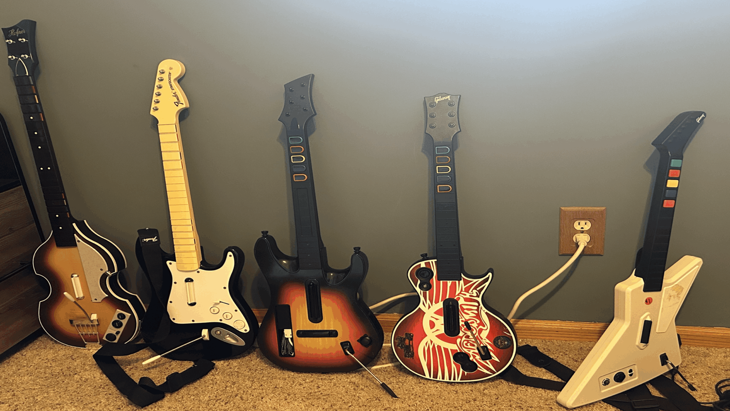 Five video game guitar controllers, a striking array of gaming accessories in different designs, are lined up against a gray wall next to a two-outlet wall socket on a carpeted floor.