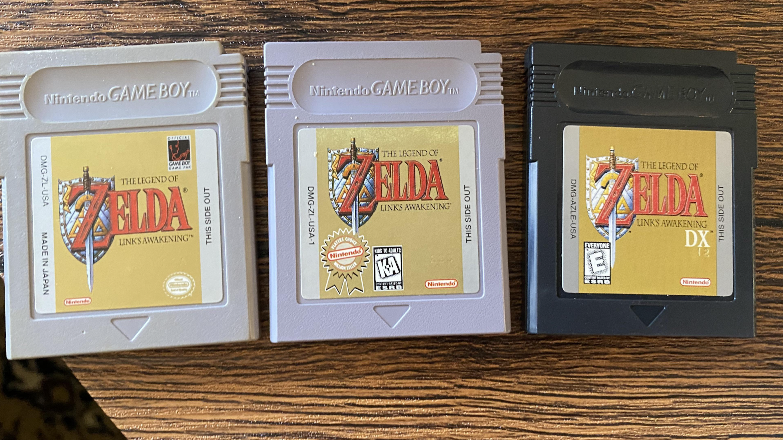 Three Nintendo Game Boy cartridges of “The Legend of Zelda: Link’s Awakening” games are displayed side by side on a wooden surface, featuring two gray and one black cartridge—a classic lineup for any fan of Zelda games.