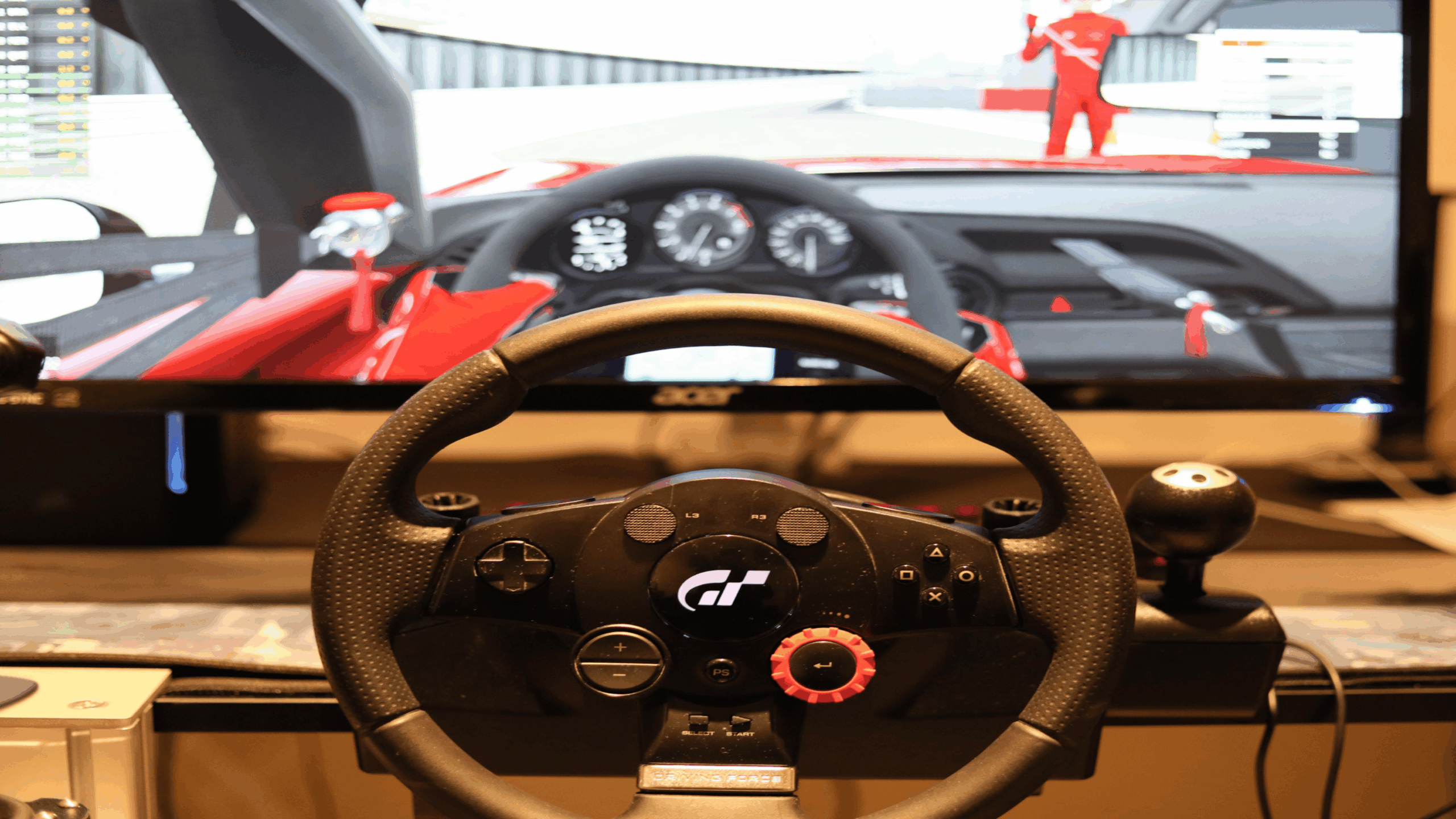 A racing game is displayed on a monitor, viewed from behind a racing wheel controller with paddle shifters and a gear stick setup, surrounded by premium gaming accessories that enhance the immersive car driving experience.
