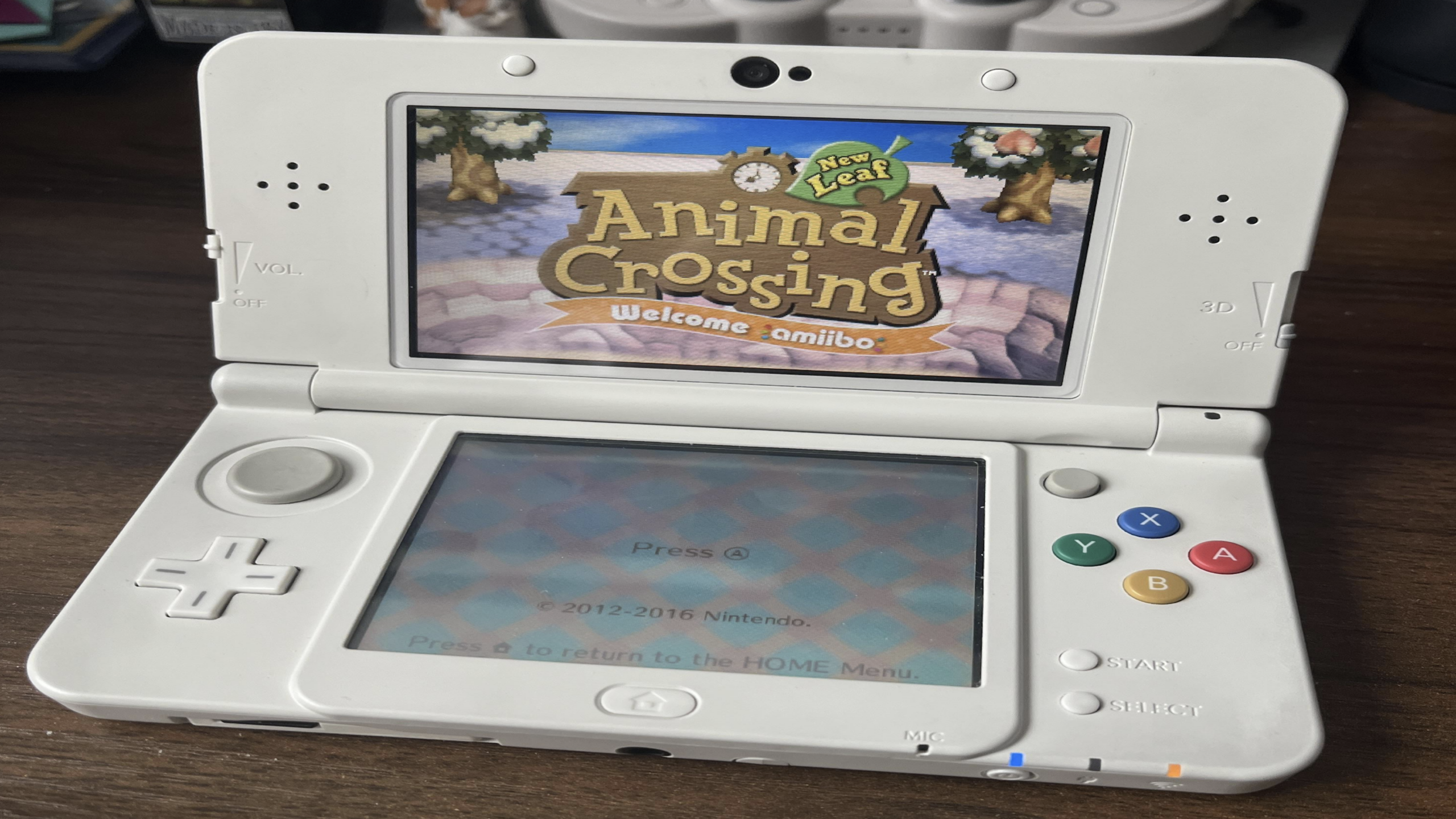 A white Nintendo 3DS console, one of the beloved Nintendo consoles, displays the "Animal Crossing: New Leaf Welcome amiibo" title screen. The console rests on a dark surface with shelves and other items blurred in the background.