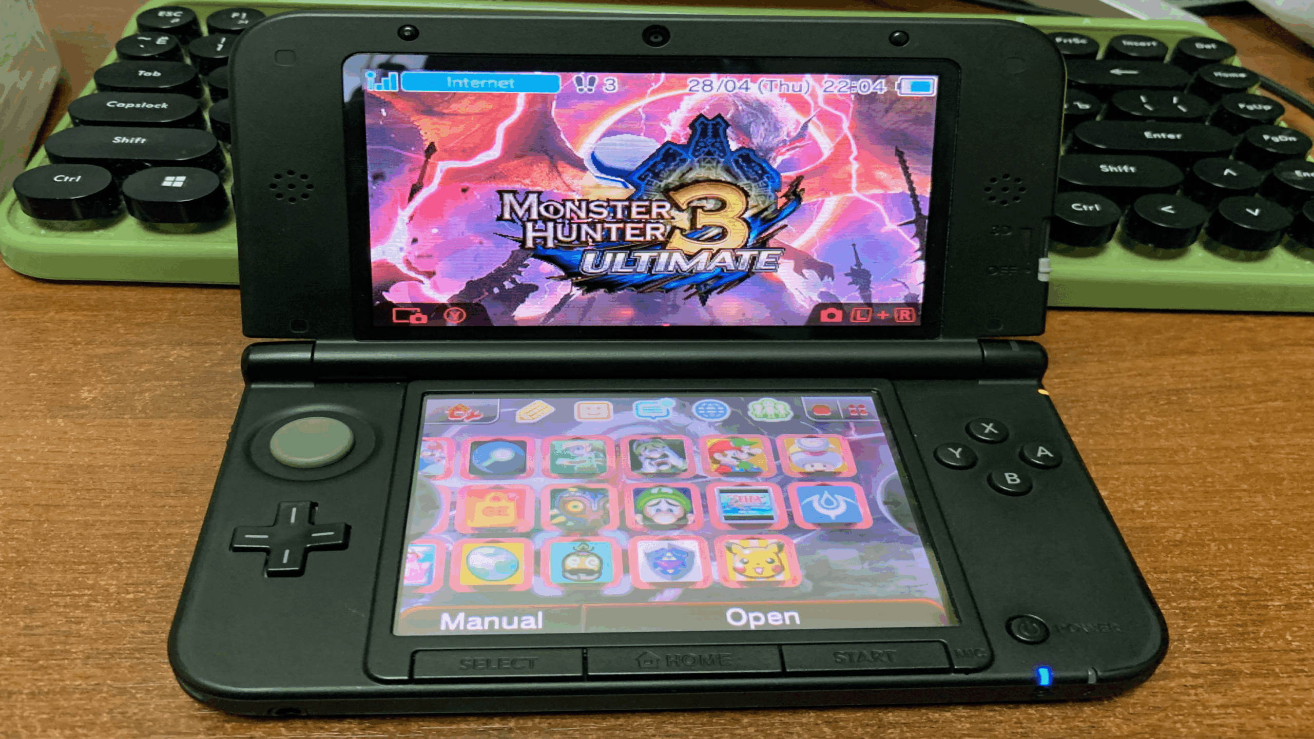 A Nintendo 3DS XL, one of the standout Nintendo Consoles, displays the game "Monster Hunter 3 Ultimate" on the top screen, with various game icons visible on the bottom screen. The console is on a wooden desk next to a green-black keyboard.