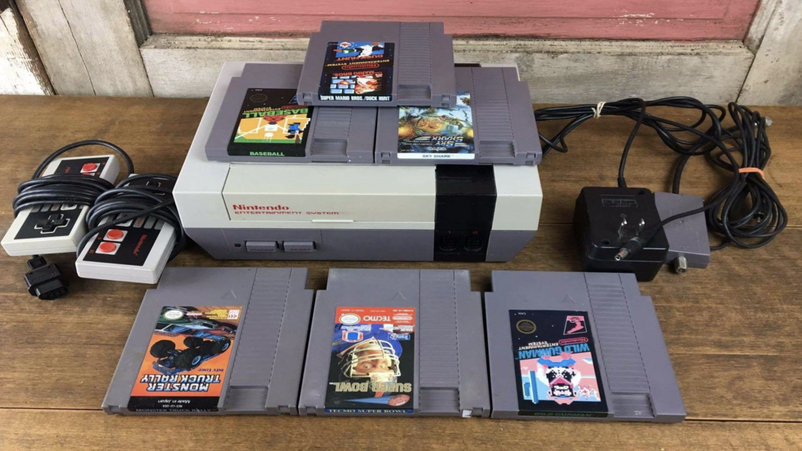 A classic piece among Nintendo Consoles, this Nintendo Entertainment System comes with two controllers, a power adapter, and six game cartridges, all displayed on a wooden table against a rustic background.