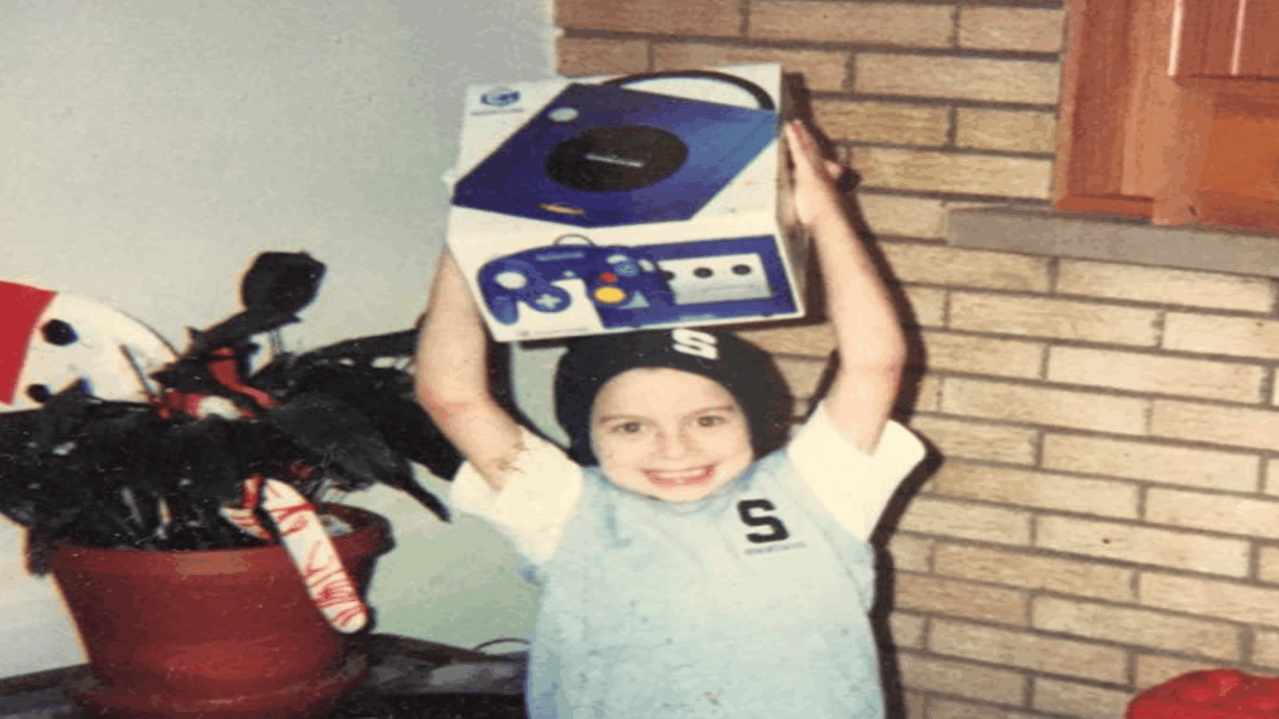 A smiling child wearing a beanie and "S" shirt excitedly holds a boxed Nintendo GameCube—one of the iconic Nintendo consoles—above their head, standing indoors near a potted plant and brick wall.