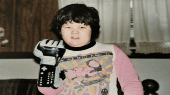 A child wearing a vintage Nintendo Power Glove&mdash;one of the most iconic gaming accessories&mdash;and a colorful graphic T-shirt stands indoors, looking at the camera. Drapes and a window are visible in the background.
