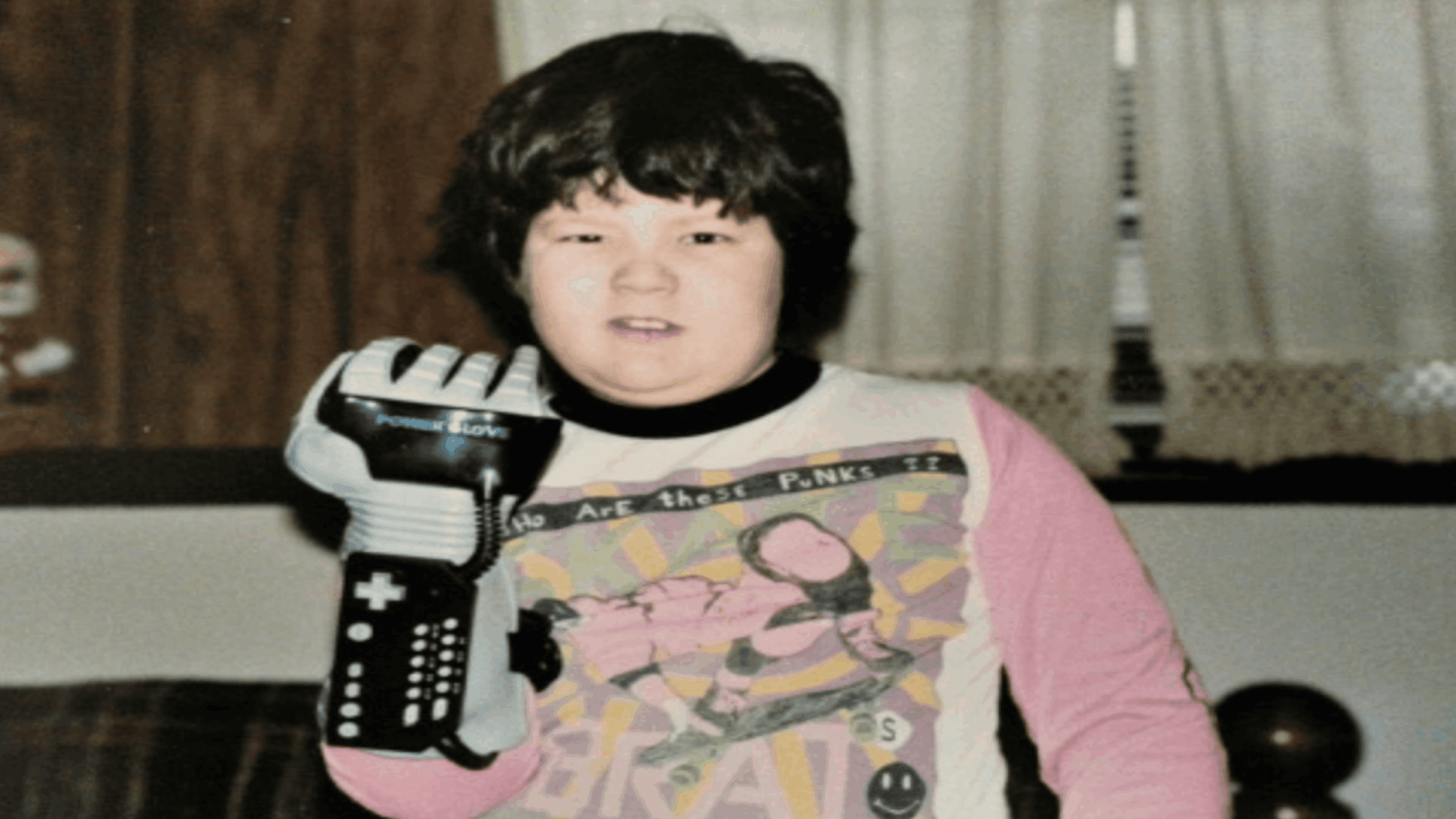 A child wearing a vintage Nintendo Power Glove—one of the most iconic gaming accessories—and a colorful graphic T-shirt stands indoors, looking at the camera. Drapes and a window are visible in the background.