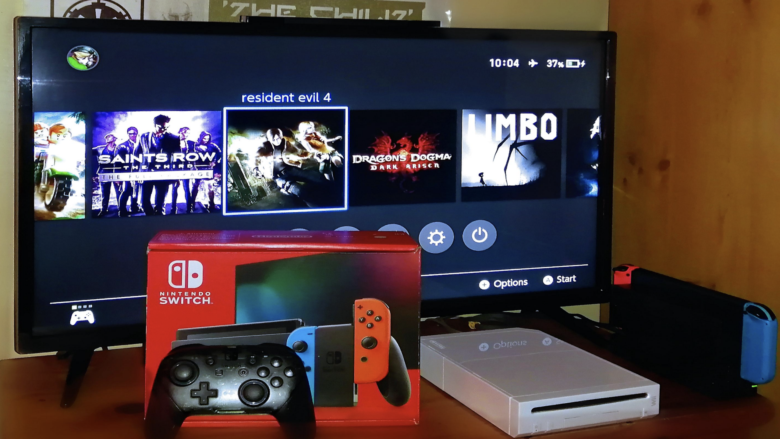 A TV displays a Nintendo Switch menu with games. In front are various consoles, including a Switch box, a black controller, a white Wii console, and the Switch dock with red and blue Joy-Cons.