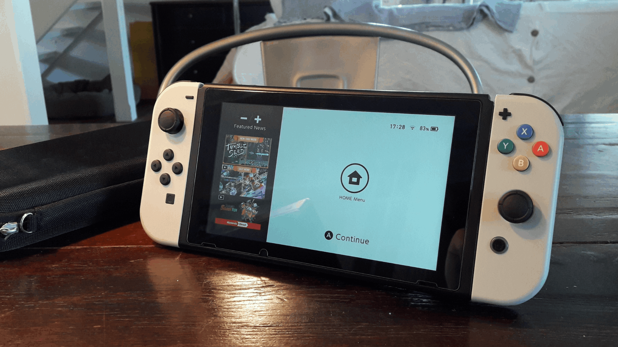 A Nintendo Switch, one of the most popular Nintendo consoles, with white Joy-Cons sits on a wooden table, displaying the Home Menu on its screen. A black carrying case is next to it, and a metal chair is in the background.