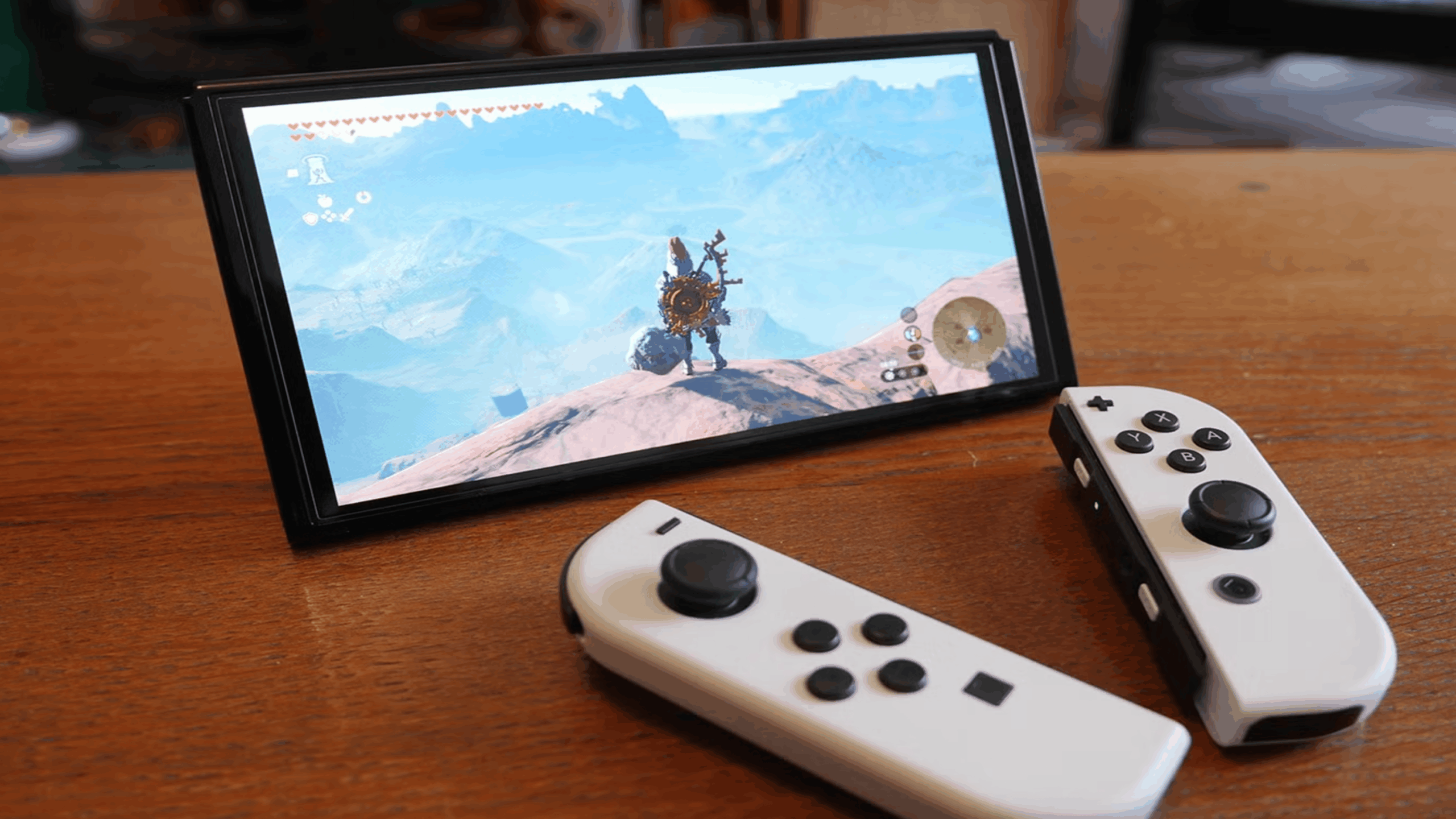 A Nintendo Switch, one of the standout Nintendo consoles, with white Joy-Con controllers rests on a wooden table. The screen displays a scene from "The Legend of Zelda: Breath of the Wild," showing a character standing on a mountain overlooking a vast landscape.