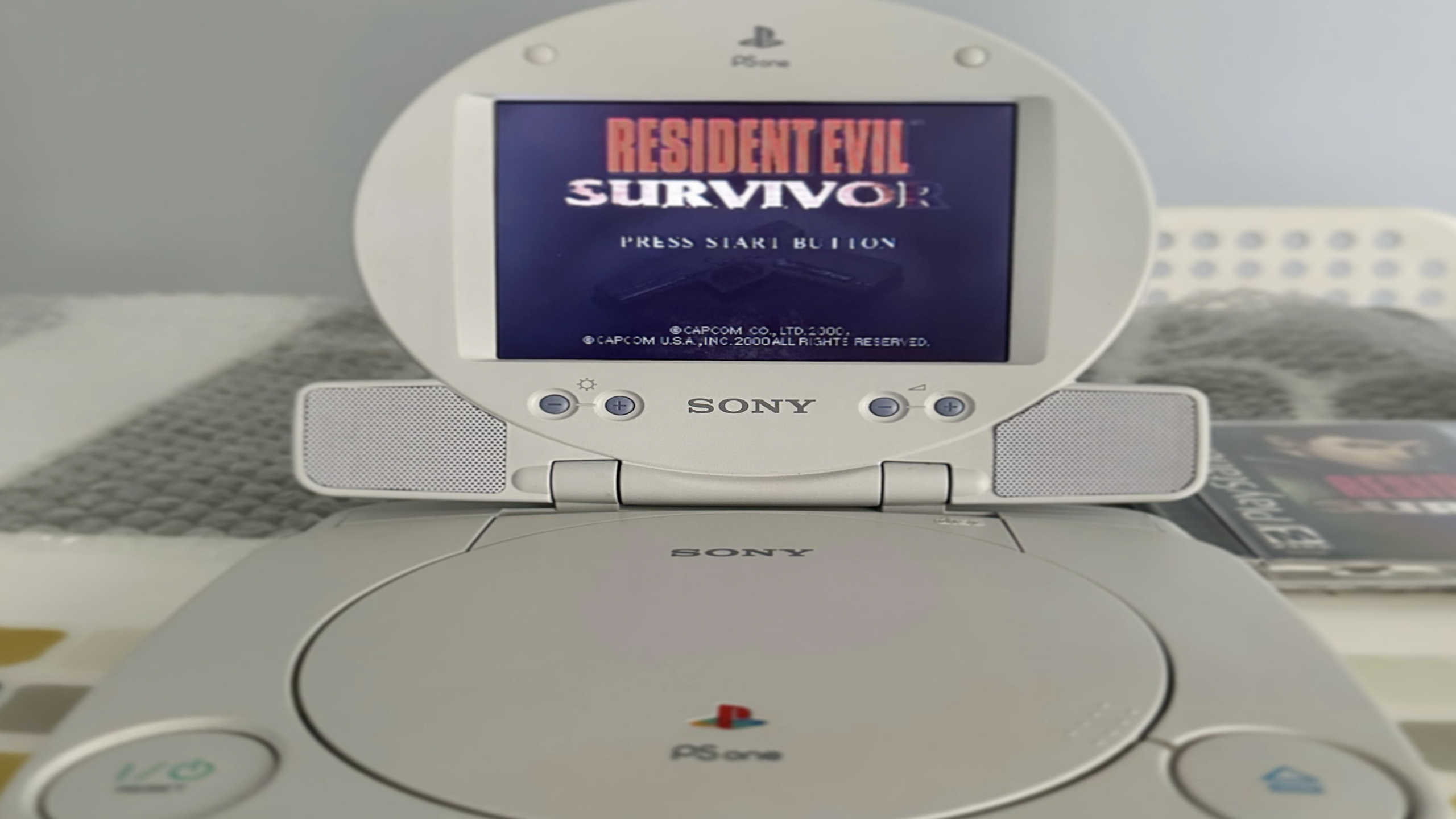 A Sony PlayStation PSone with an attached LCD screen displays the title screen for “Resident Evil: Survivor.” The console and screen are on a table with some blurred items in the background.