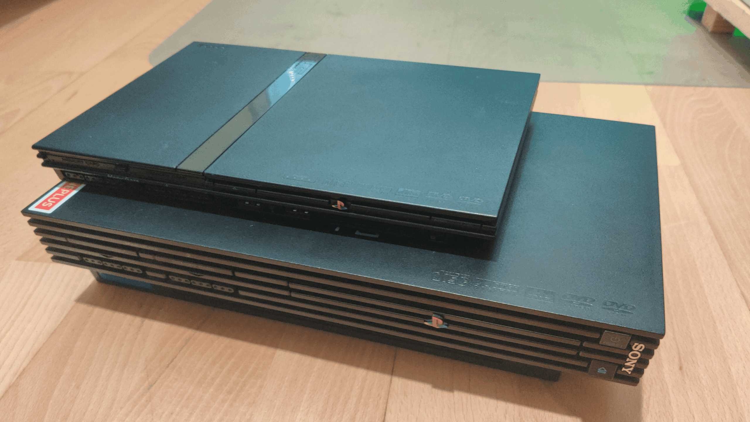 Two Sony PlayStation 2 consoles, one slim model stacked on top of the larger original PlayStation model, are placed on a wooden floor.