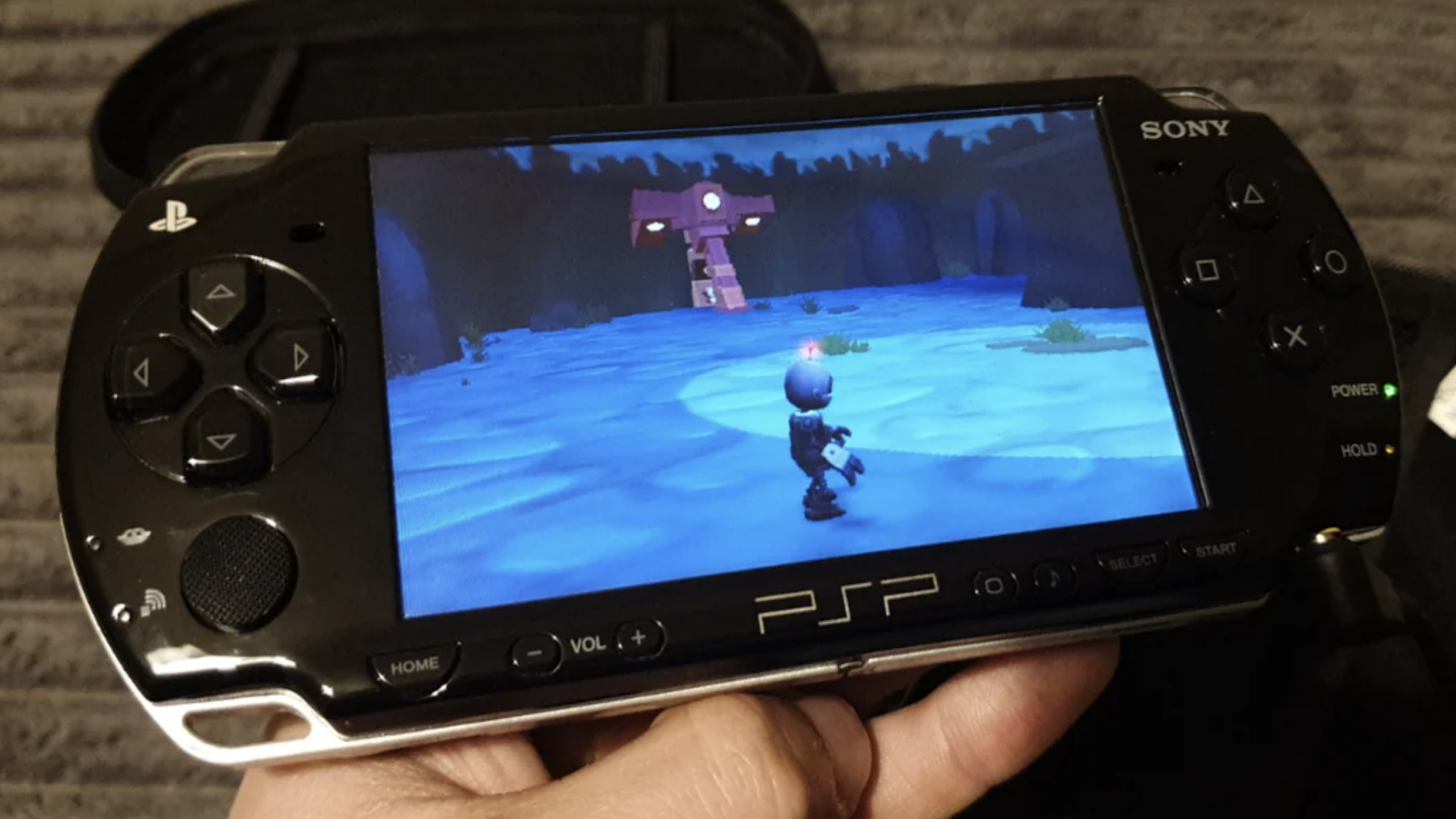 A hand holds a Sony PlayStation PSP displaying a video game with a small character on a blue landscape, facing a structure with stairs and lights.