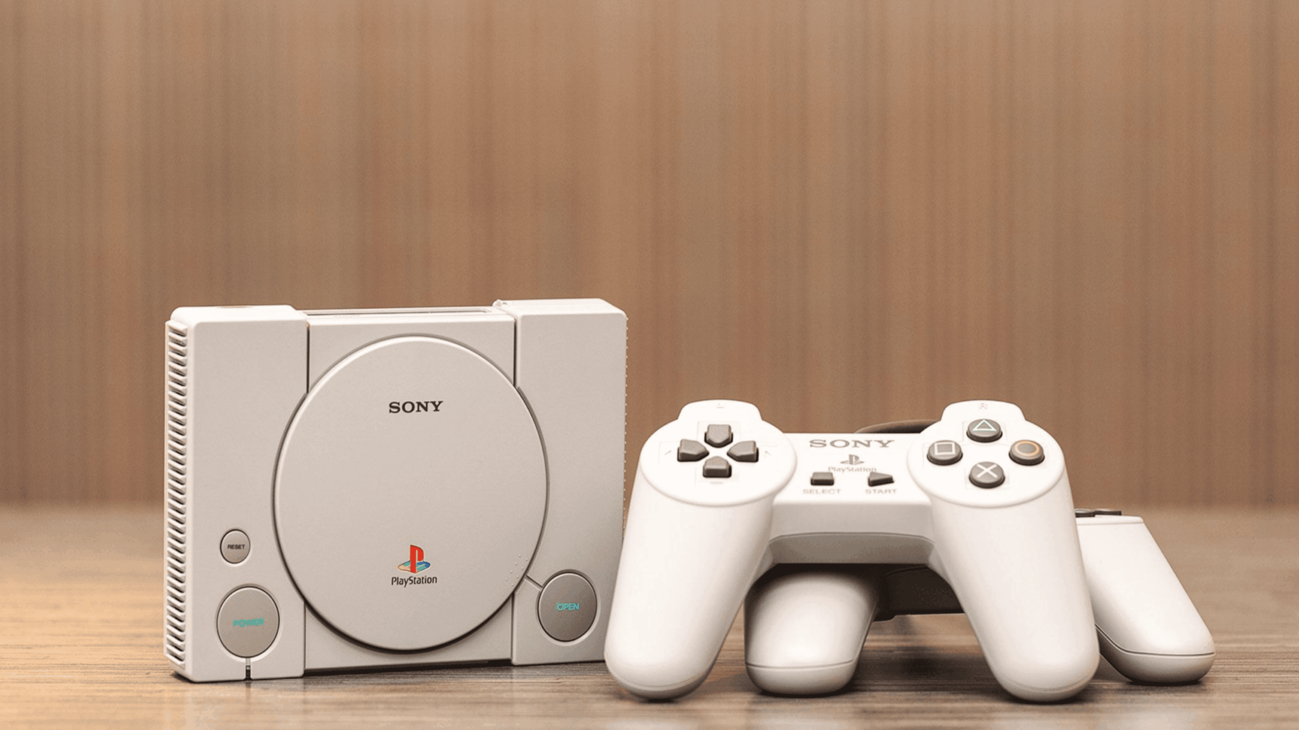 A Sony PlayStation Classic console sits on a wooden surface with a matching PlayStation controller beside it, both in light gray color, against a blurred beige background.