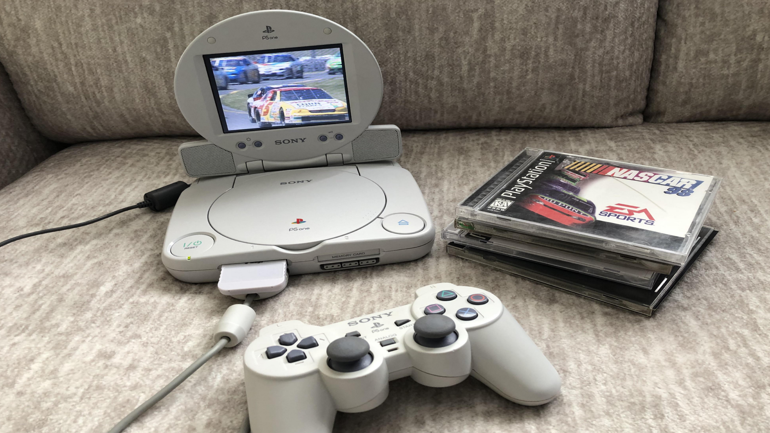 A white PlayStation One console with attached screen displays a racing game. A controller, one of several essential gaming accessories, is connected, and several PlayStation game cases, including NASCAR 98, are stacked nearby on a beige couch.
