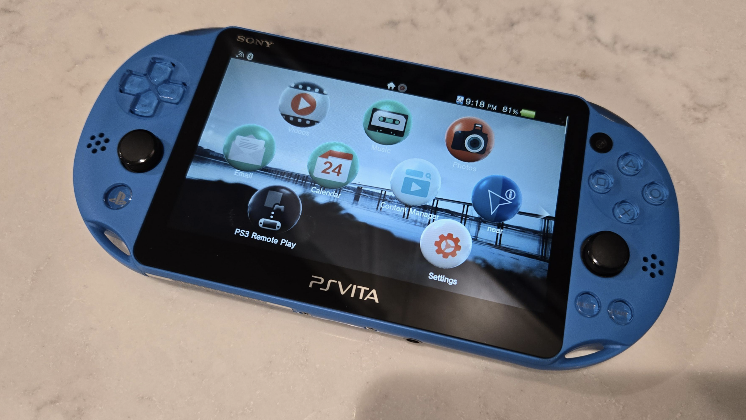 A blue Sony PS Vita PlayStation handheld gaming console displays its home screen with various app icons, lying on a light-colored surface.