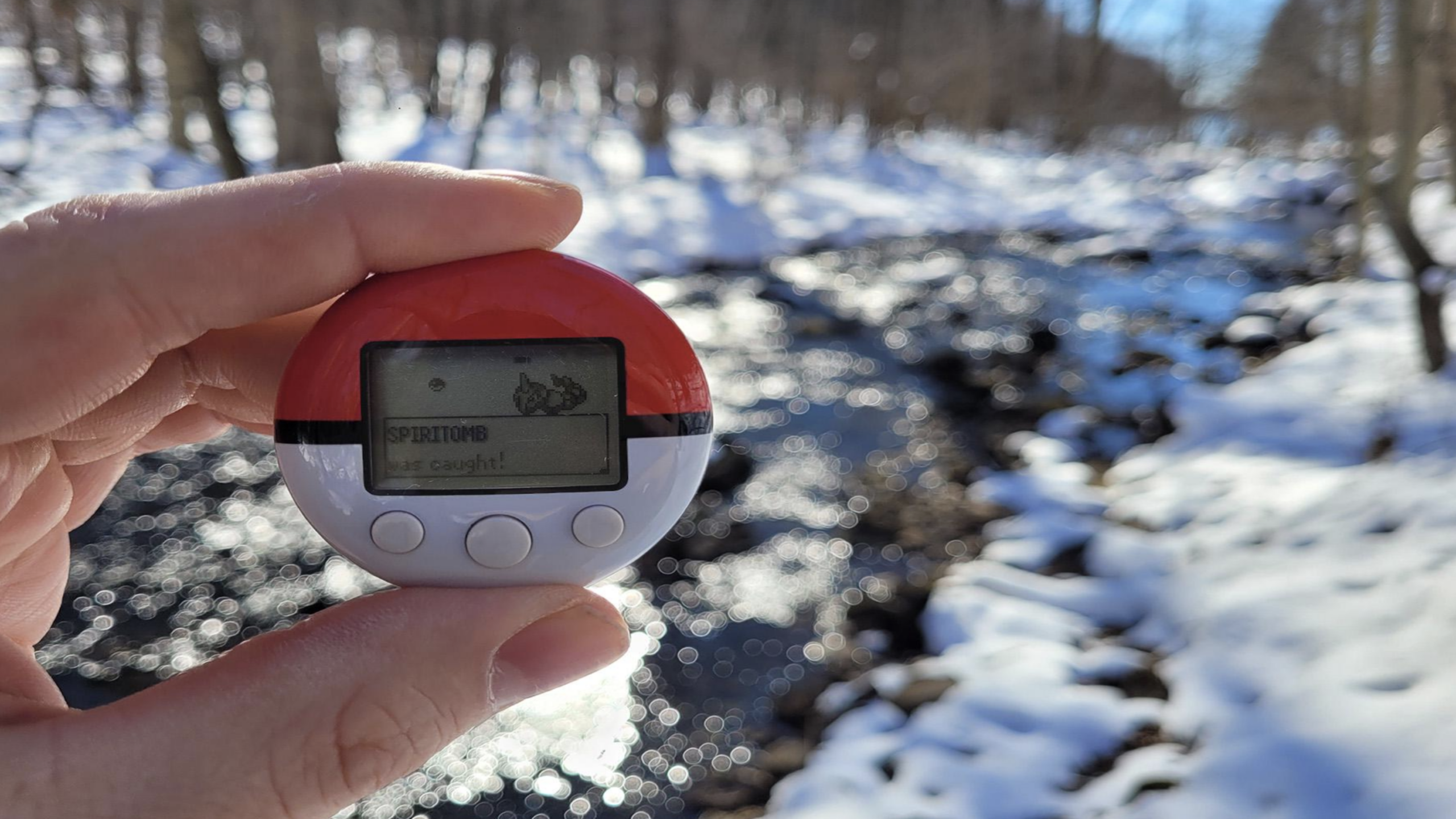 A hand holds a Pokéwalker, one of the most iconic gaming accessories, displaying a captured Spiritomb near a snowy creek in a sunlit forest.
