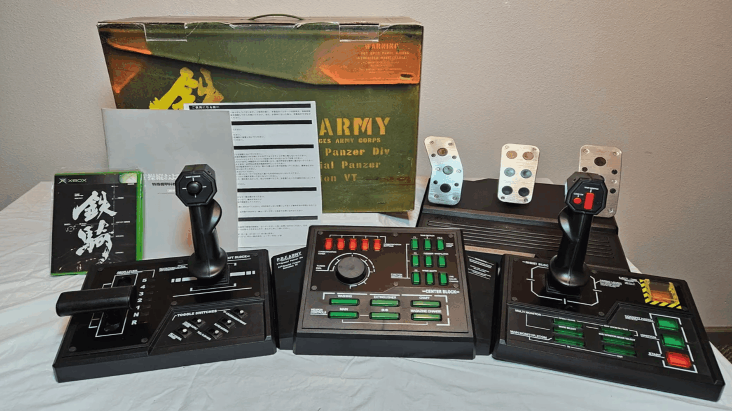 A complex game controller setup featuring three control panels, multiple buttons, two joysticks, foot pedals, manuals, and a green and brown box labeled "Steel Battalion"—all items are arranged on a white surface alongside various gaming accessories.