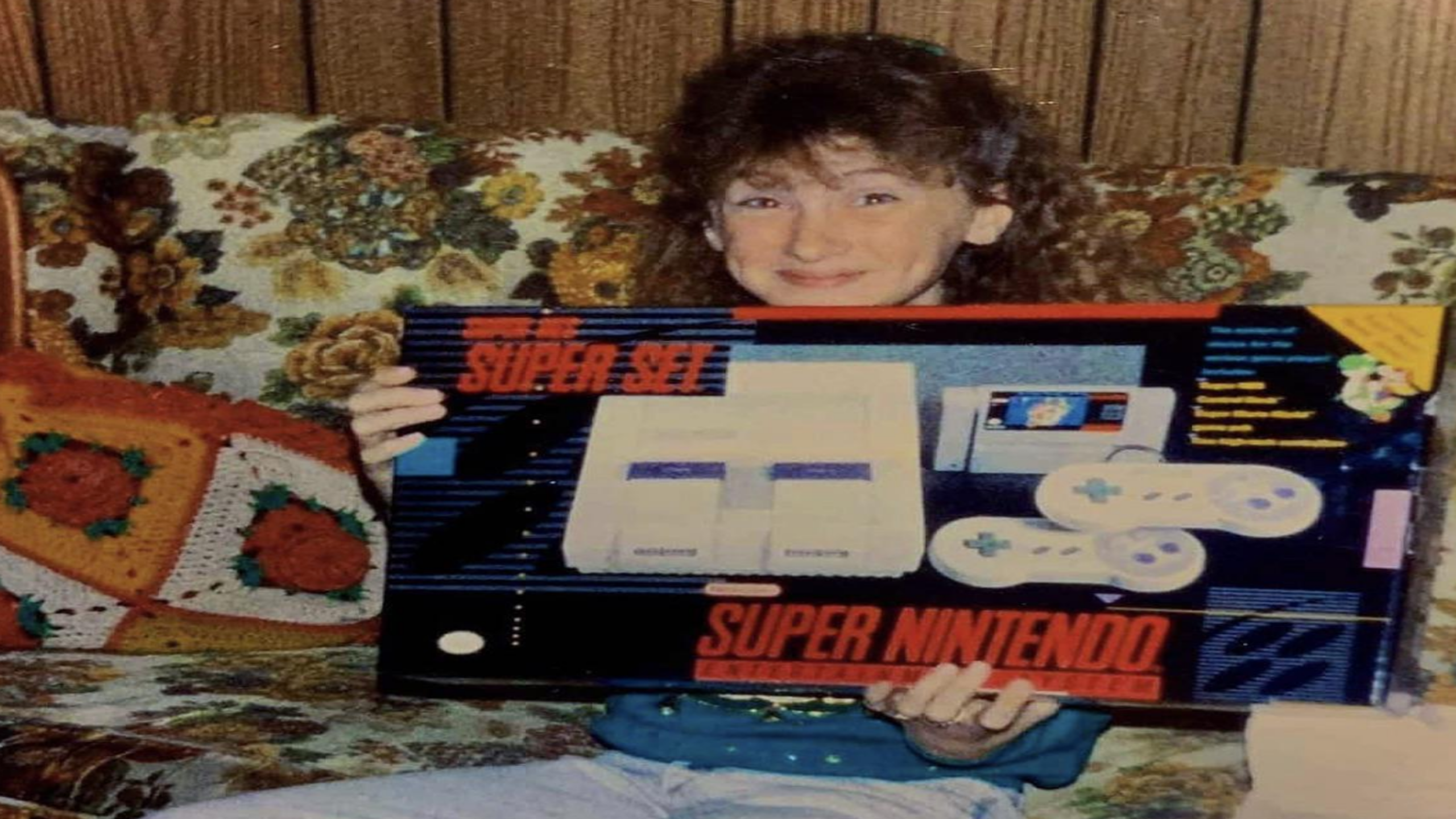 A smiling child sits on a patterned couch, holding a Super Nintendo Entertainment System box—the crown jewel among classic Nintendo consoles. The couch has a colorful crochet pillow, and a wood-paneled wall is in the background.