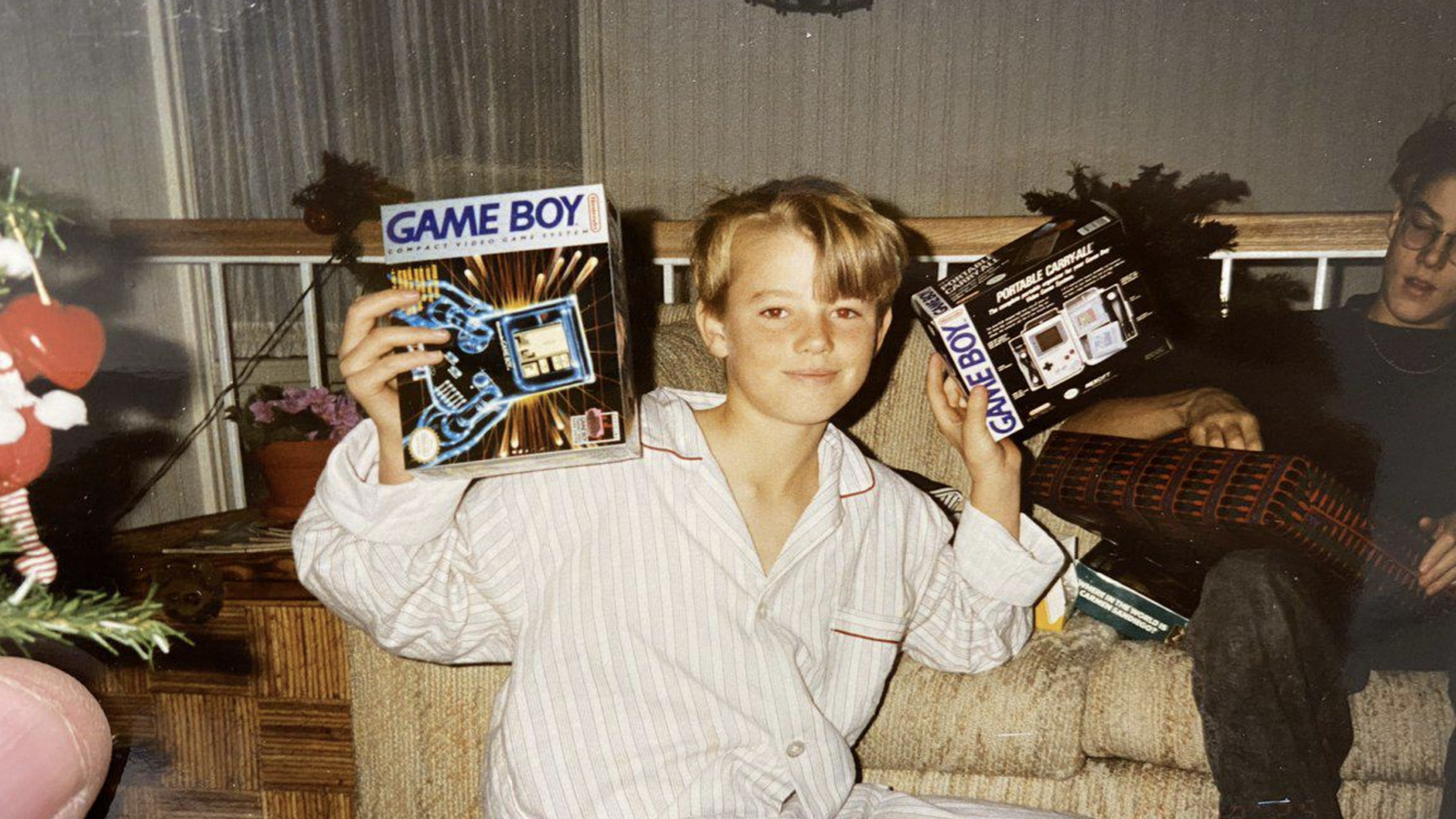 A smiling child in pajamas sits on a couch, proudly holding up a Game Boy console and a Tetris package. Holiday decorations and another person are visible in the background.