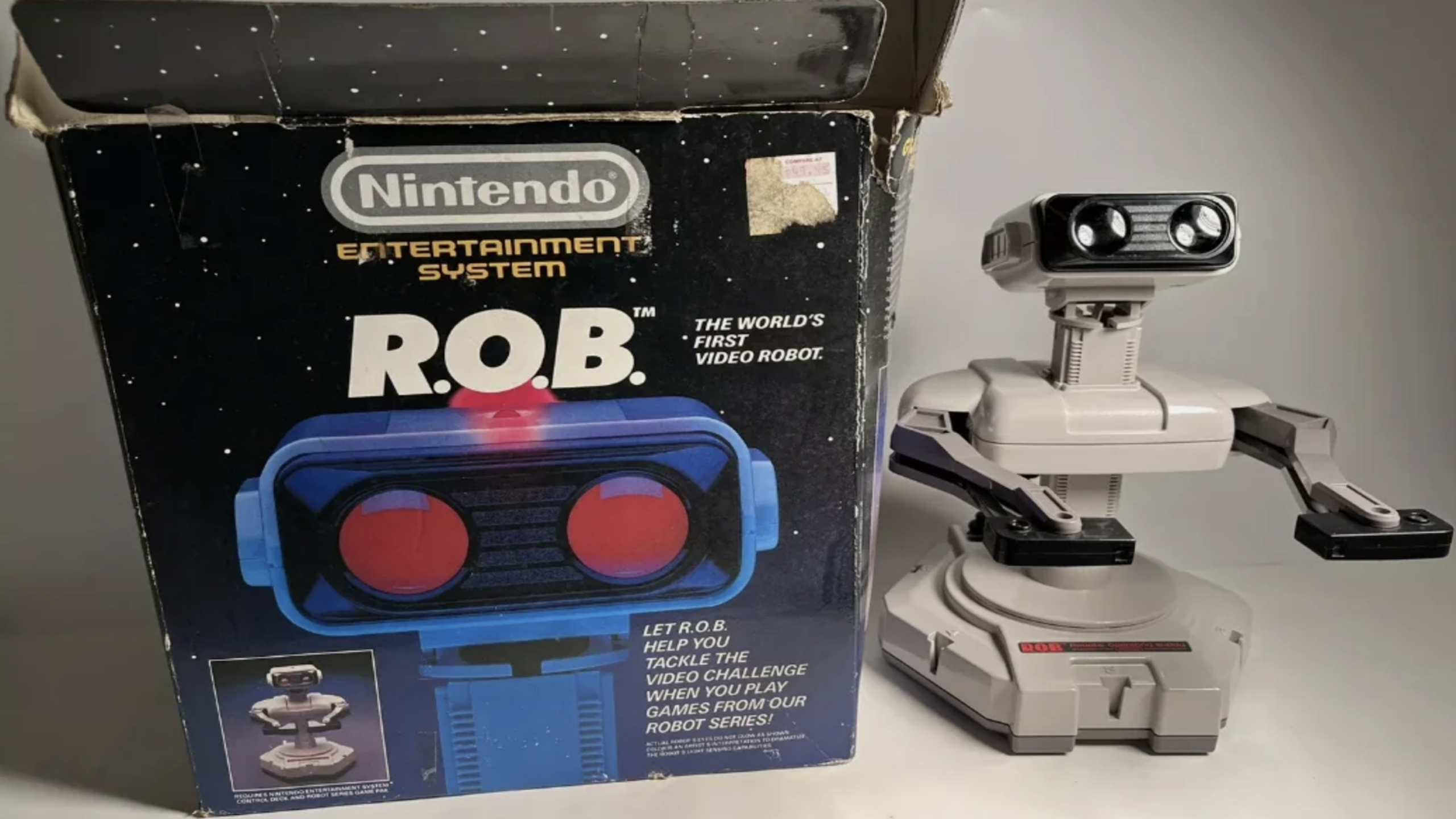A Nintendo R.O.B. robot, one of the most iconic gaming accessories of its era, stands next to its original box, which features branding and an image of the robot’s face with glowing red eyes.