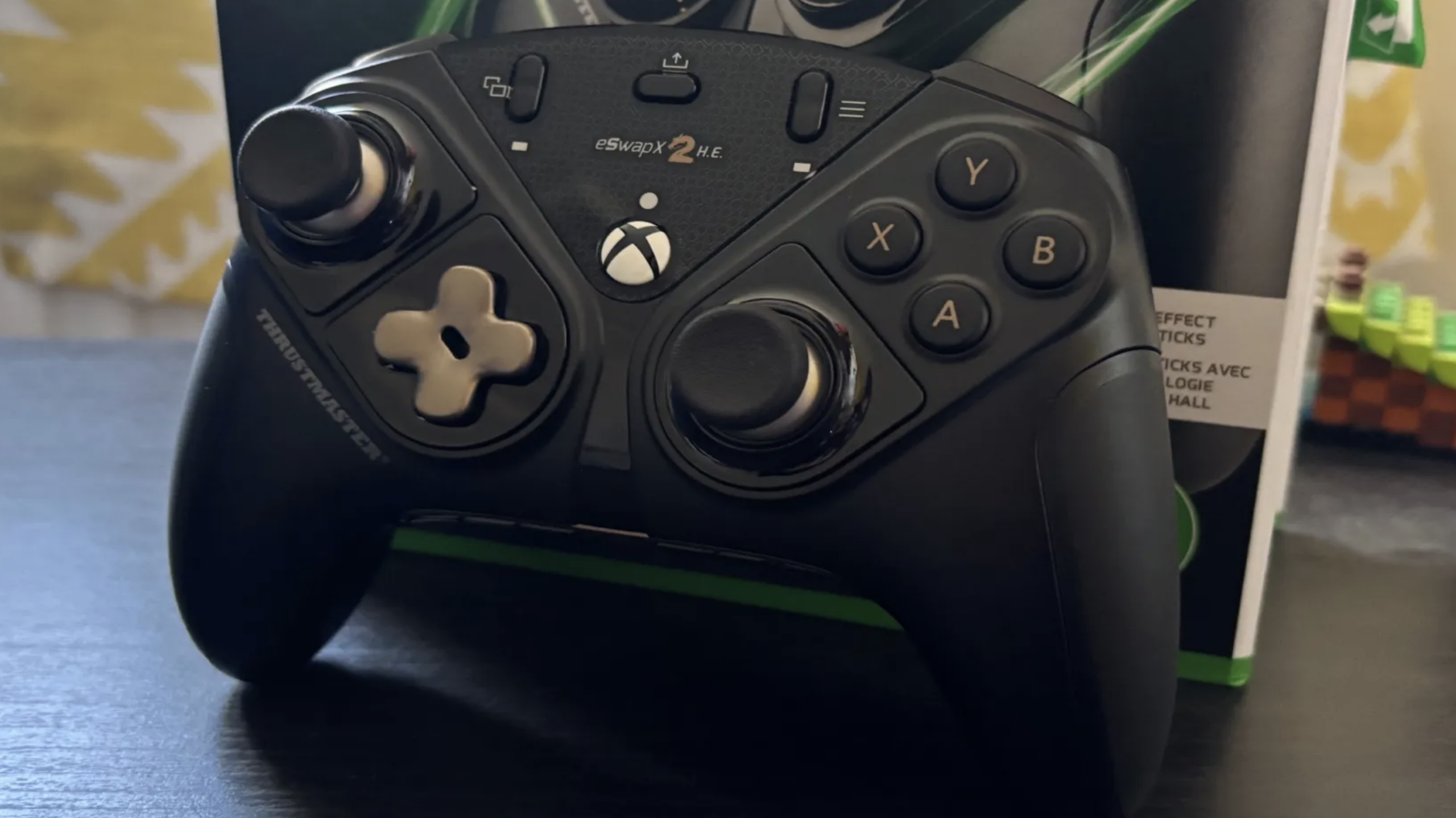 A black Thrustmaster eSwap X2 Xbox controller, known for its premium and expensive gaming features, is displayed on a table with its box in the background and part of a toy visible on the right.