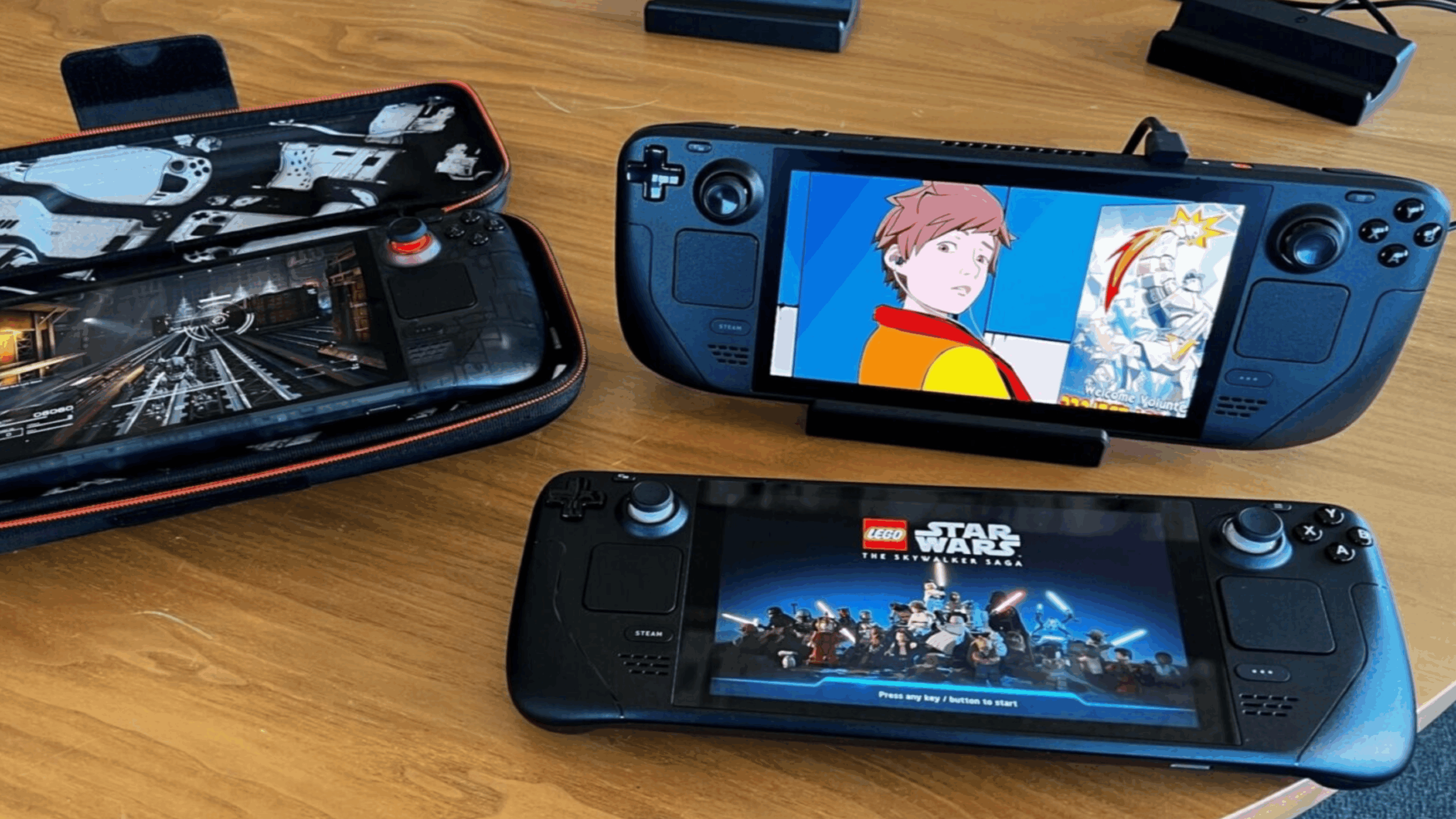 Three handheld gaming consoles are on a wooden table. One is in a protective case, while two consoles are powered on, displaying different games or shows—including LEGO Star Wars and an animated scene featuring a character in a red jacket.