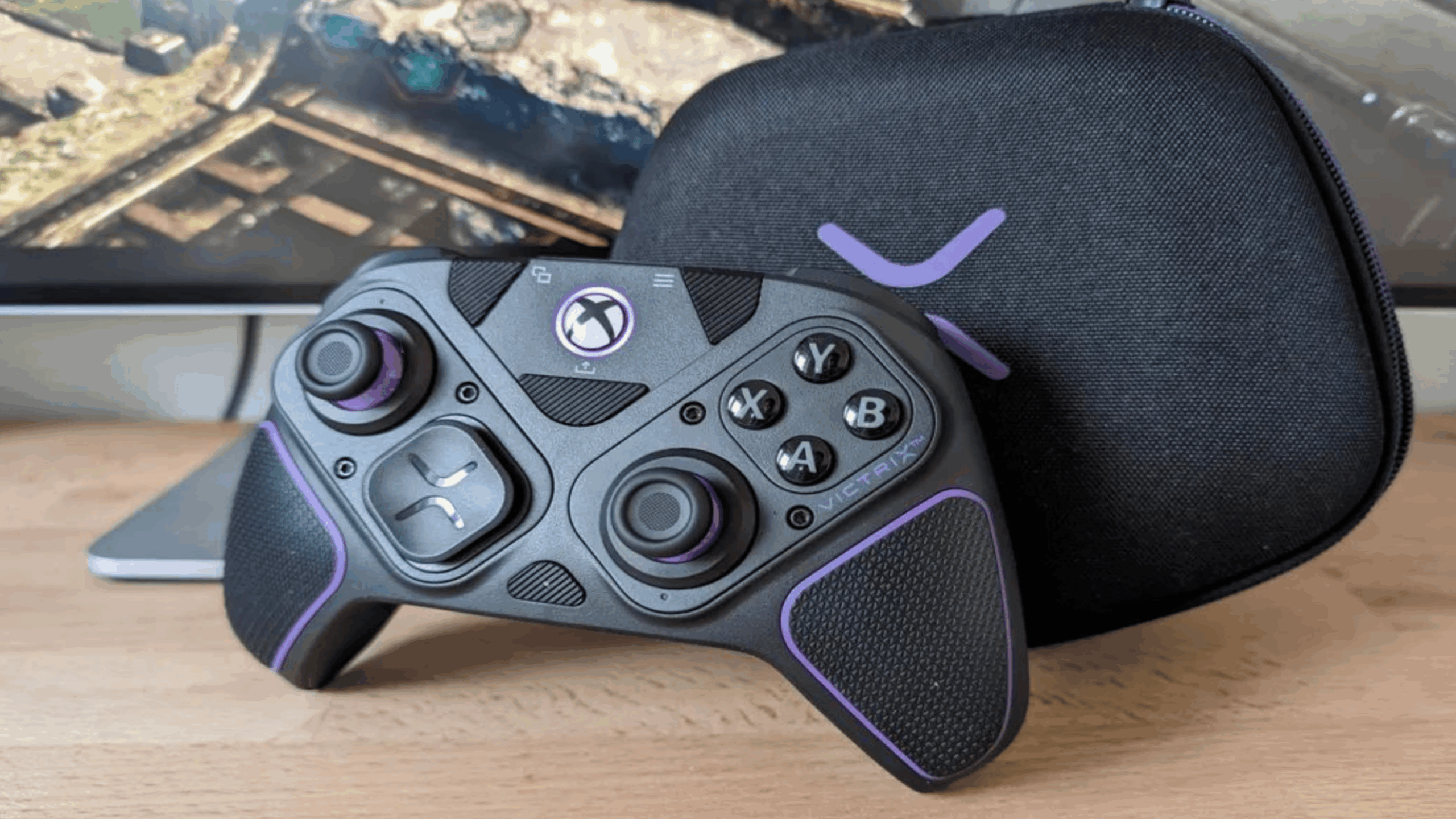 An expensive gaming controller with black and purple accents is resting in front of a black carrying case and a computer monitor on a wooden desk.