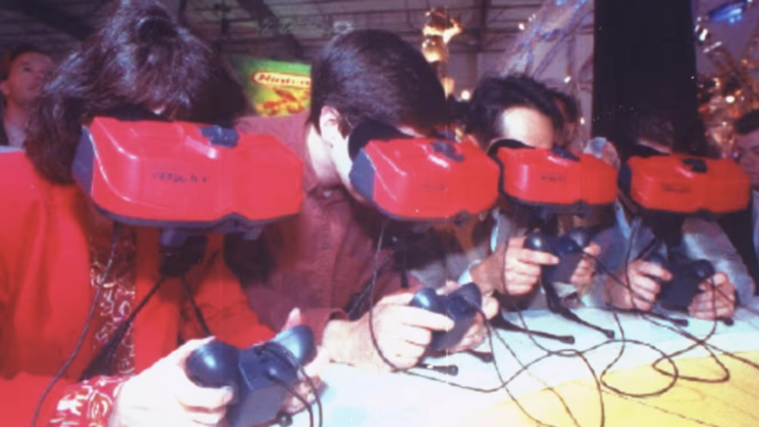 Four people sitting side by side wear large red headsets and hold controllers, appearing to test Nintendo Consoles and virtual reality gaming devices at an event or exhibition.